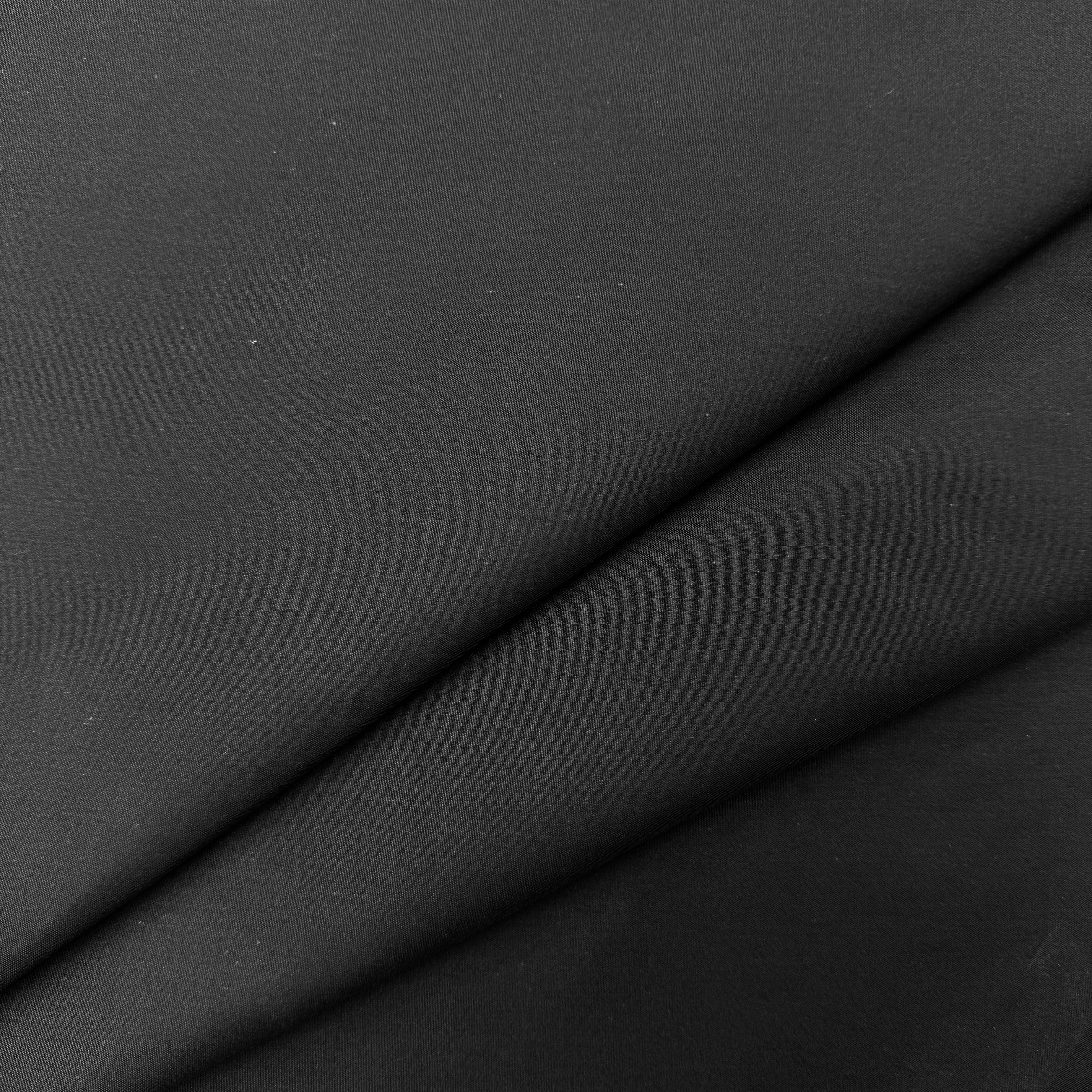 A close-up of black fabric with soft diagonal folds showing its smooth texture.