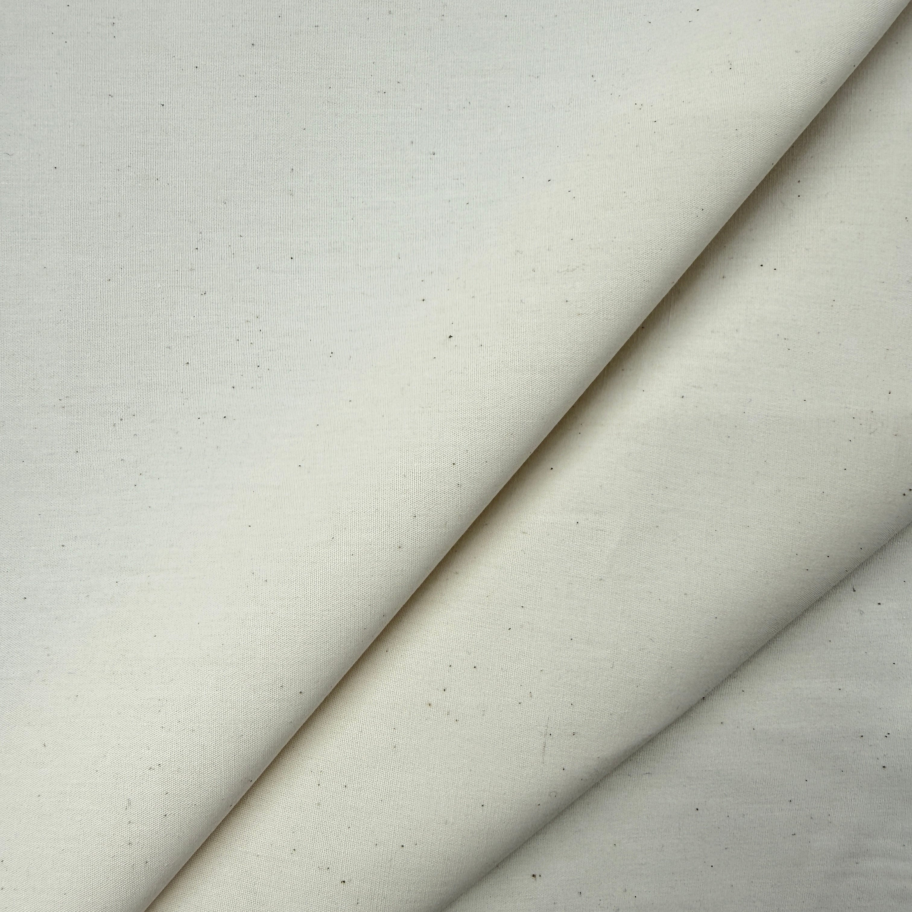 A close-up of a light warm off-white fabric with a subtle woven texture and tiny dark specks, folded diagonally across the frame.