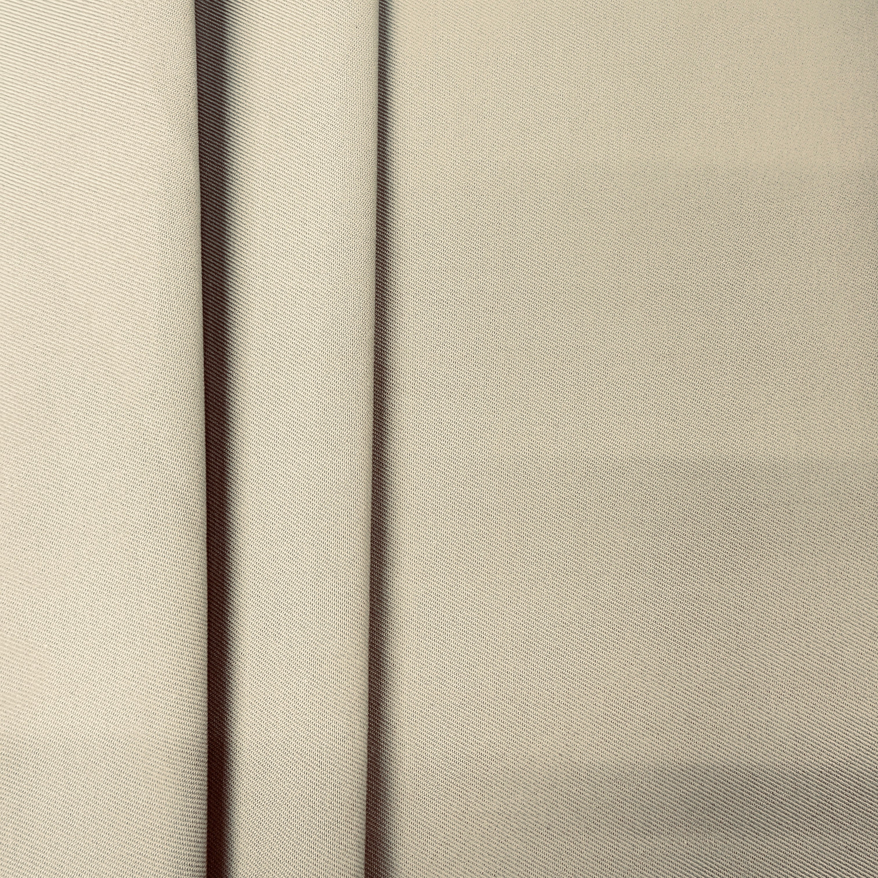 A close-up of a light beige fabric with a fine diagonal twill texture, showing soft vertical folds.