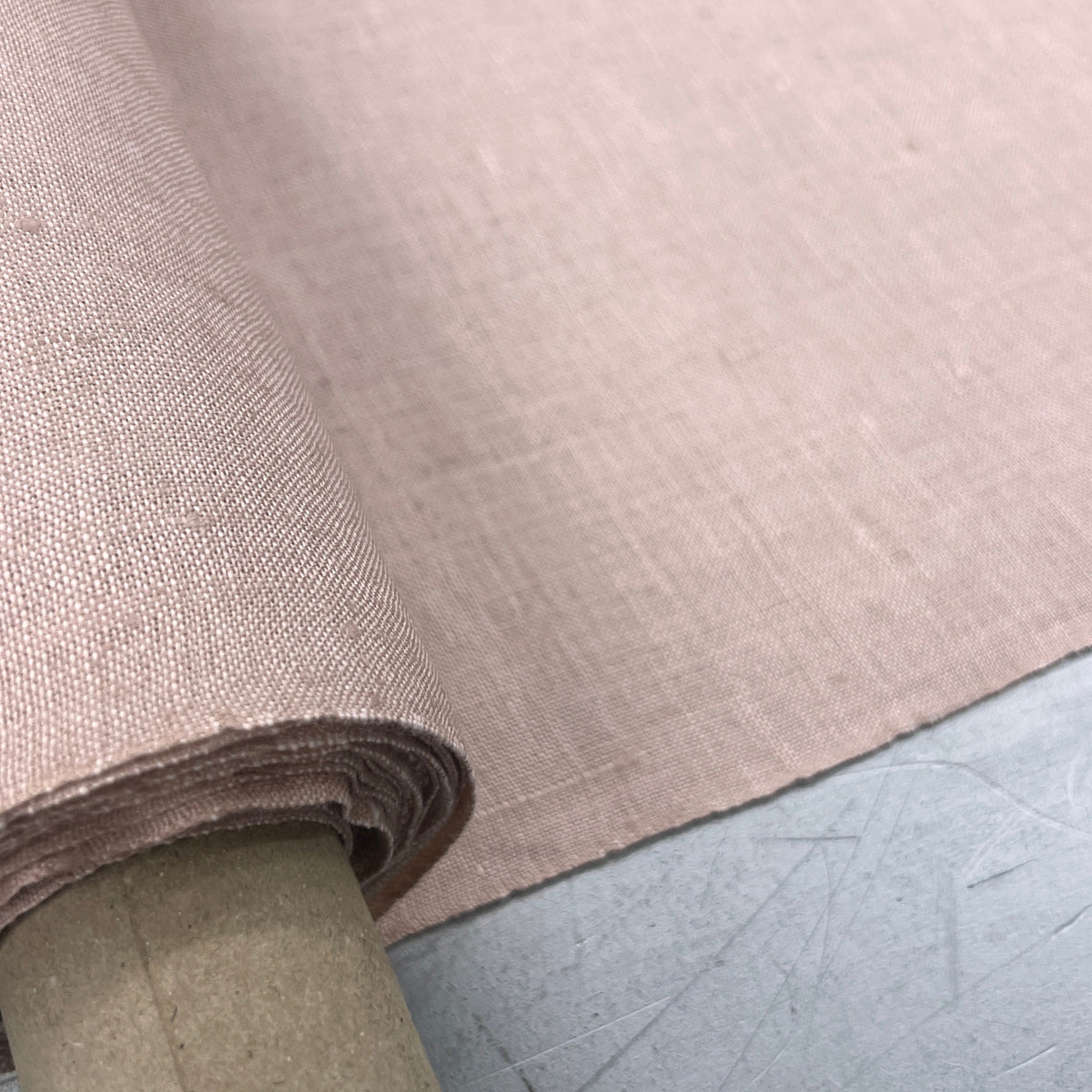 A close-up of a rolled piece of dusty pink fabric with a visible woven texture, mounted on a cardboard spool and laid over a gray surface.