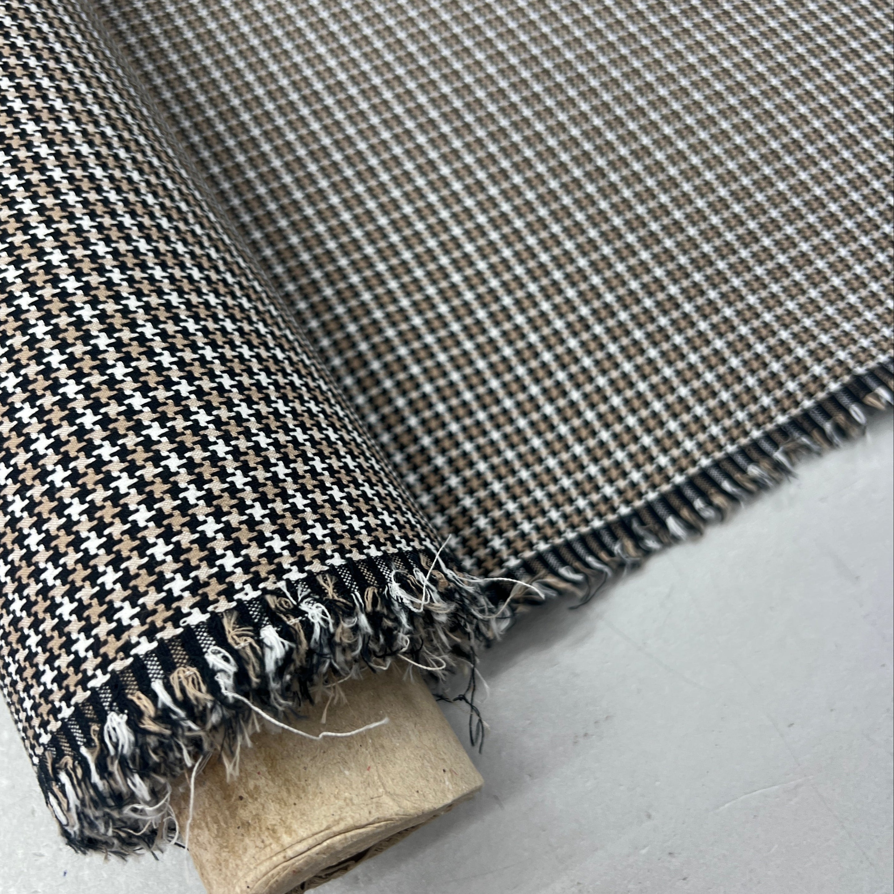 A close-up of a rolled piece of fabric with a small-scale houndstooth pattern in black, white, and beige, showing the frayed woven edges and a light-colored cardboard core.