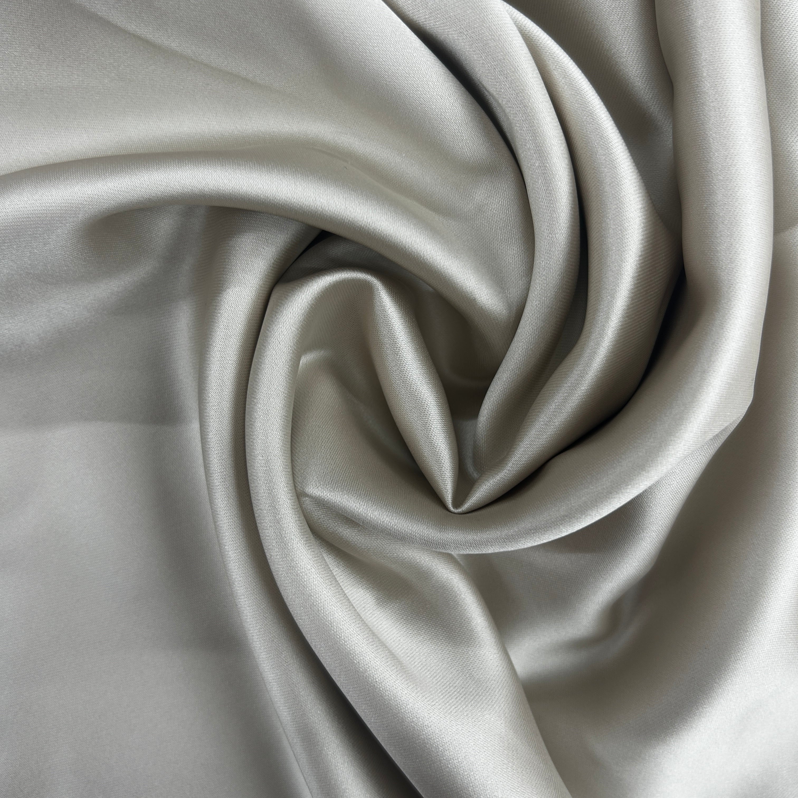 A close-up of draped satin fabric in a light silver-gray color, arranged in elegant swirls to show off its smooth texture and sheen.
