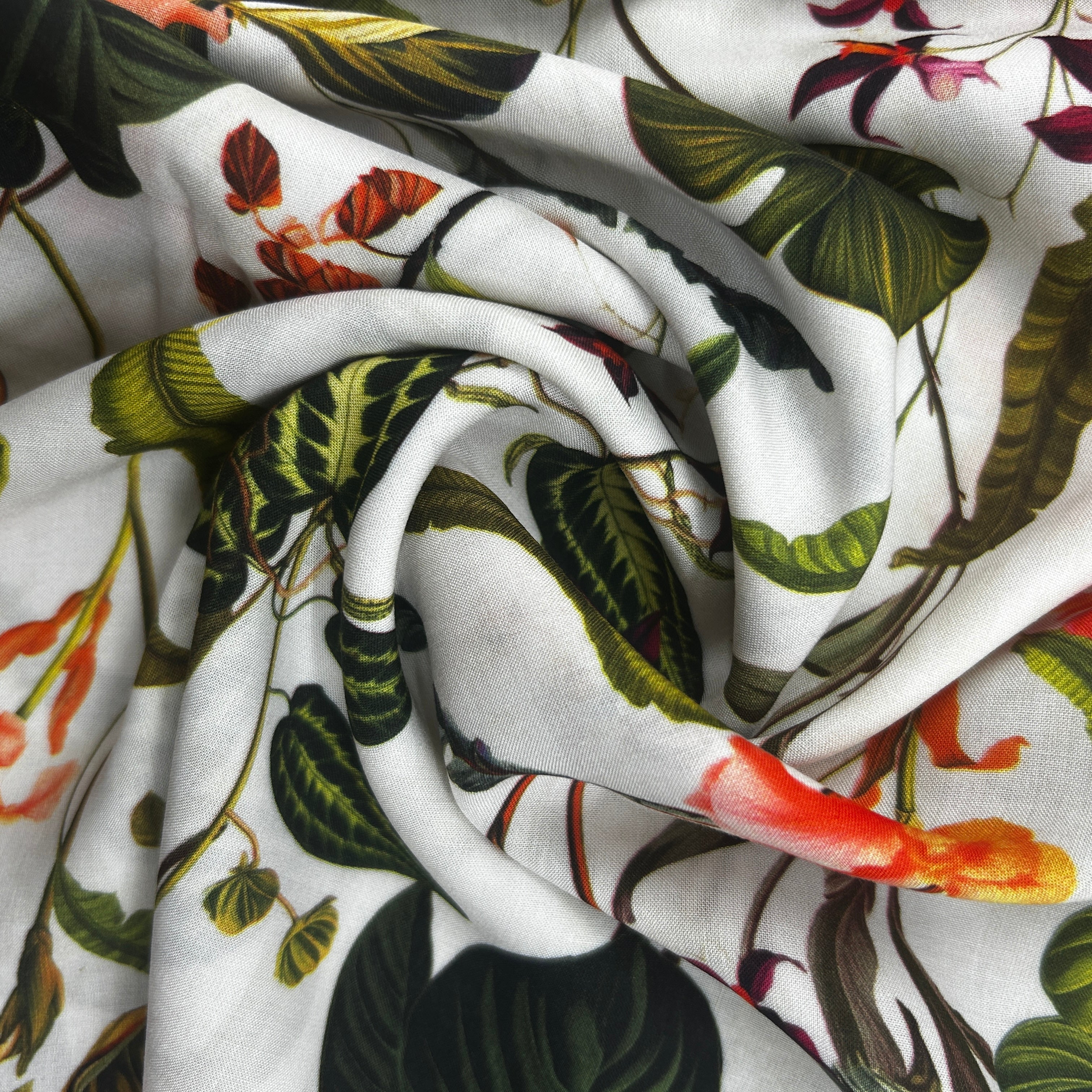 A close-up of draped white fabric printed with a tropical botanical pattern featuring green leaves, dark green foliage, and orange and red flowers.