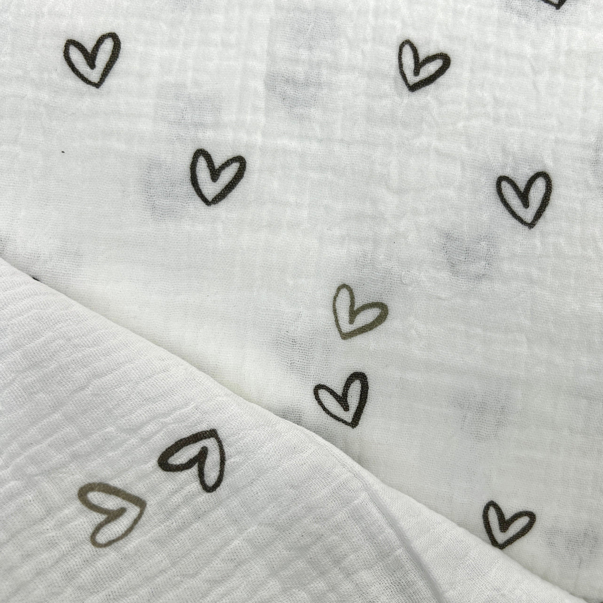 White muslin fabric with a delicate pattern of small hand-drawn hearts in black and muted olive green, folded to show its soft, textured cotton-gauze weave.
