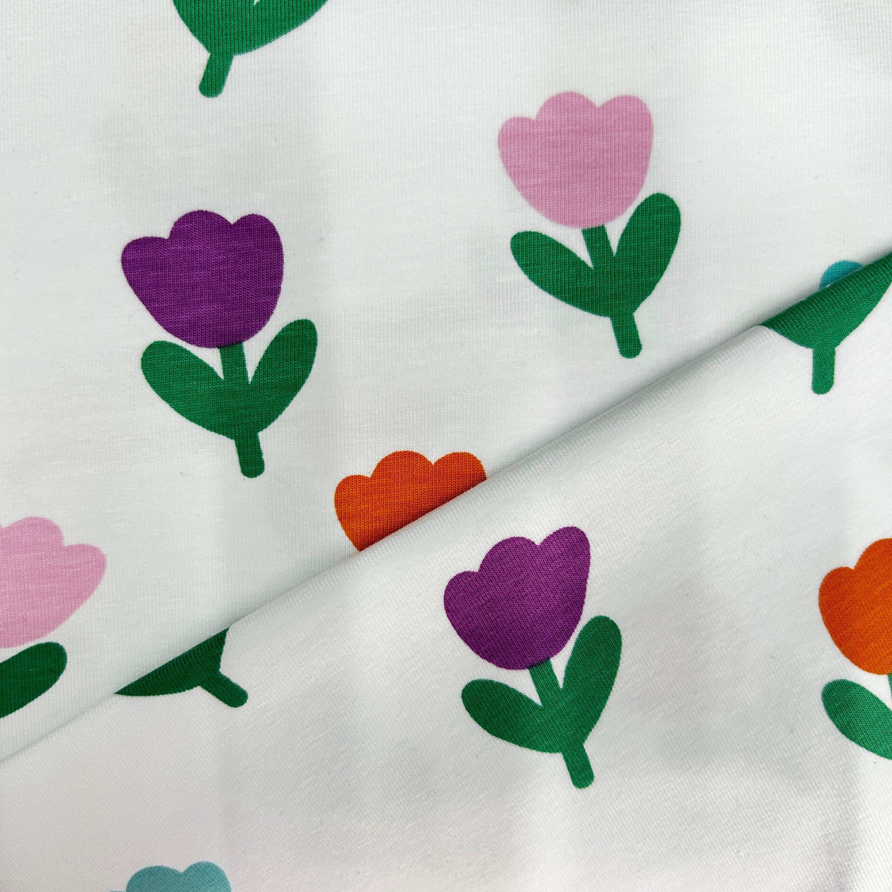White fabric with a repeating pattern of tulips in purple, pink, orange, and green, folded diagonally to show the material texture.