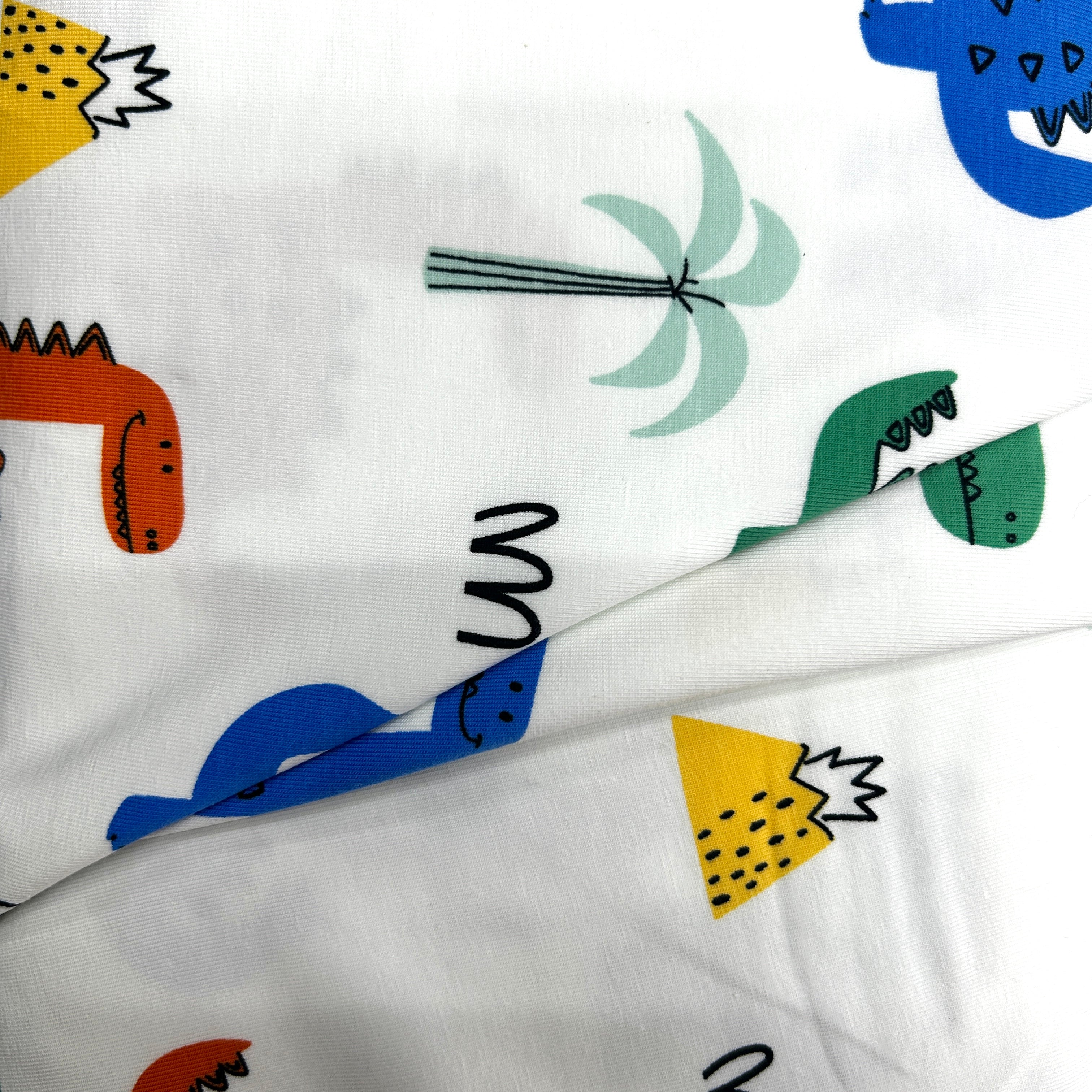 White muslin fabric with a colorful dinosaur and palm tree print, folded to show the texture and pattern details.