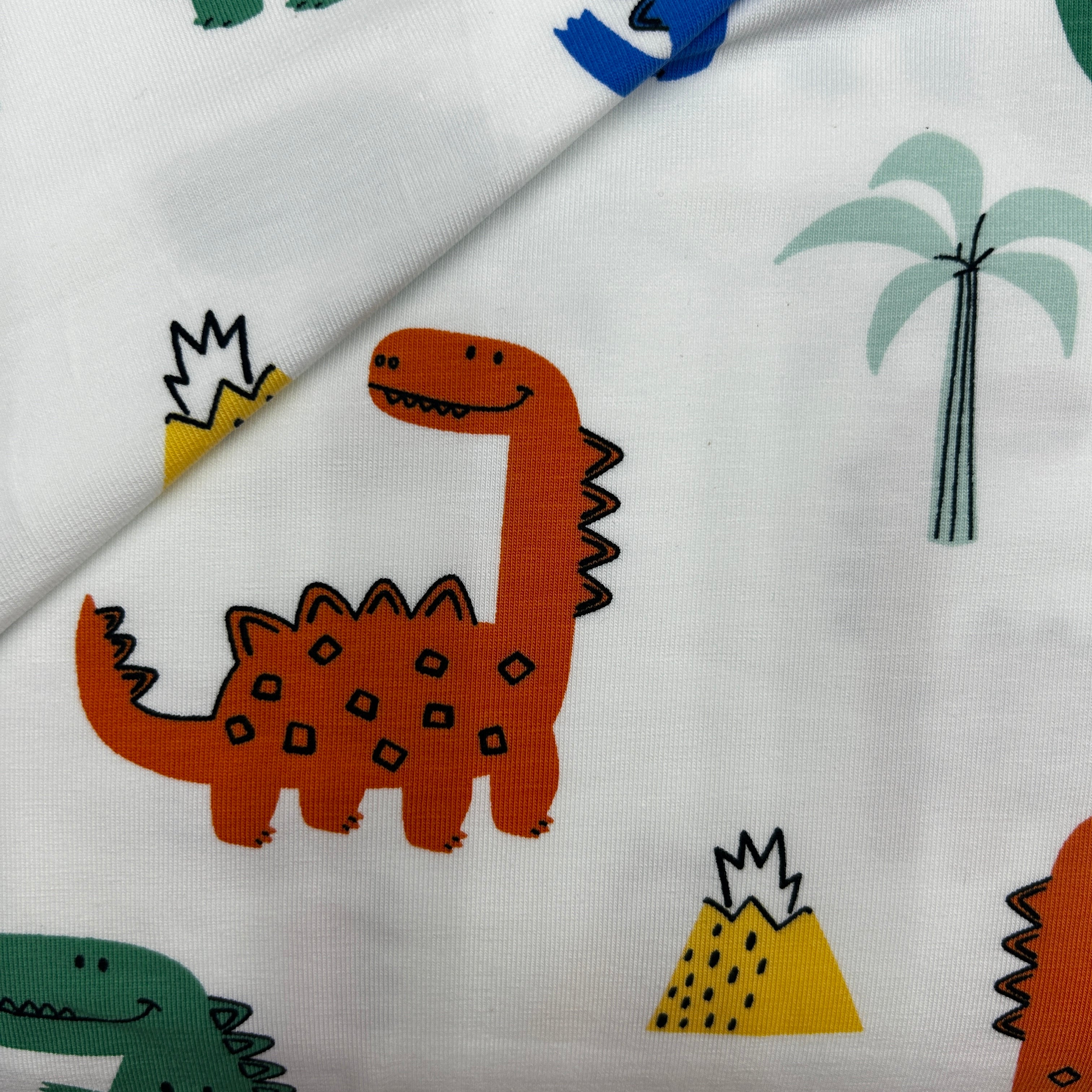 White fabric with a colorful dinosaur print, featuring a large orange-brown Stegosaurus, a green dinosaur, a yellow volcano, and a palm tree.