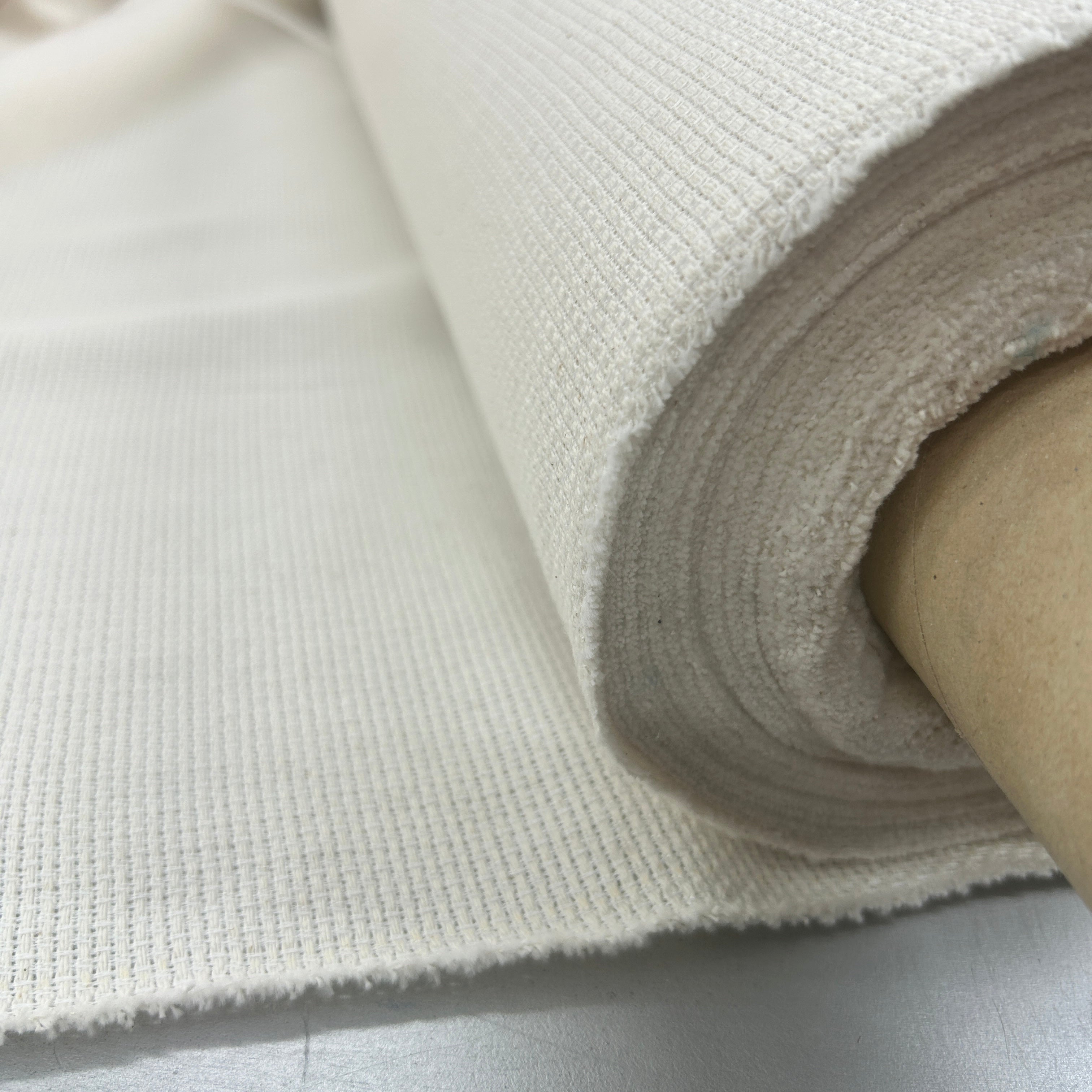A close-up of a rolled off-white woven fabric or canvas material on a light gray surface, showing the textured weave and tan cardboard core.