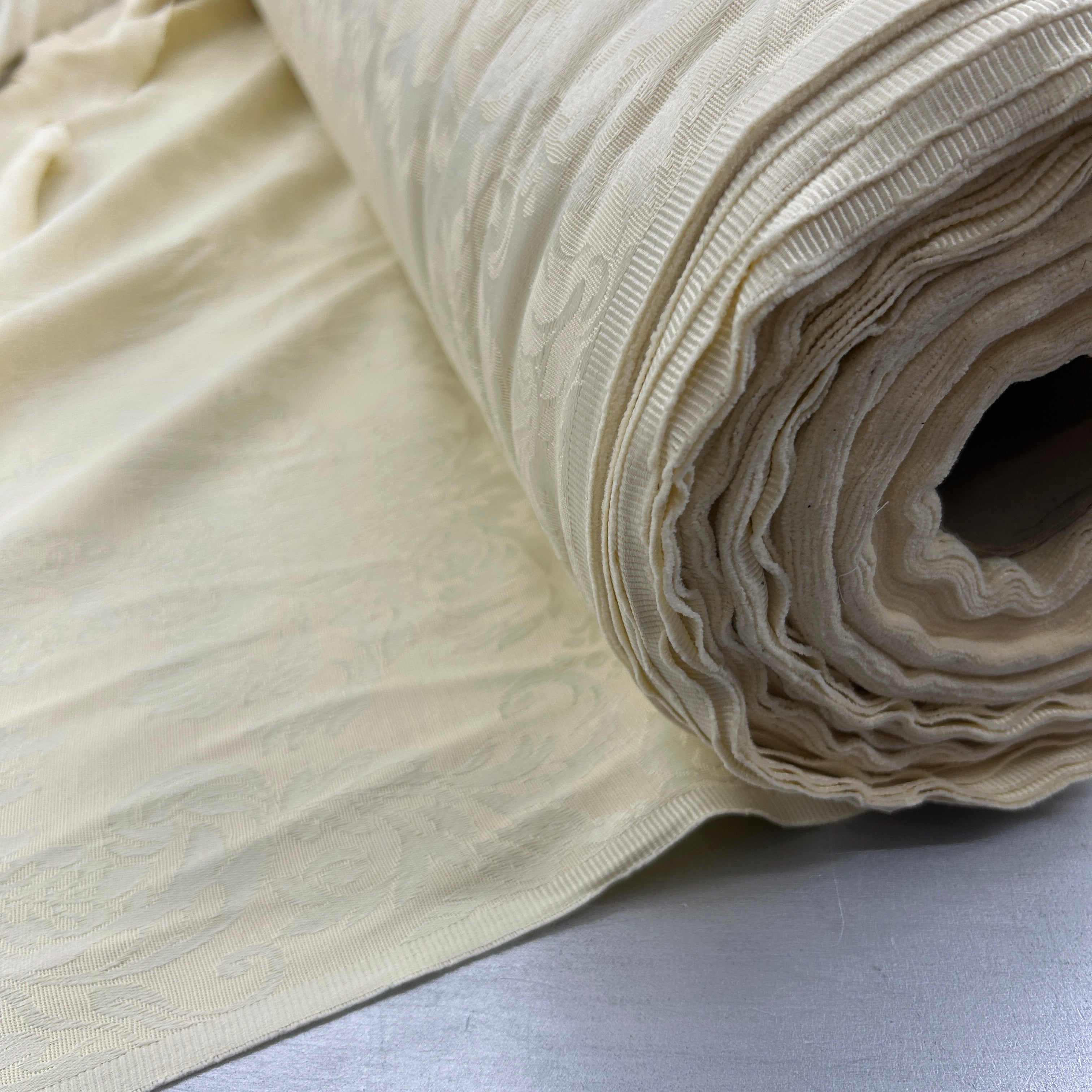 A close-up of a rolled cream-colored fabric with a subtle woven damask pattern, resting on a light gray surface.