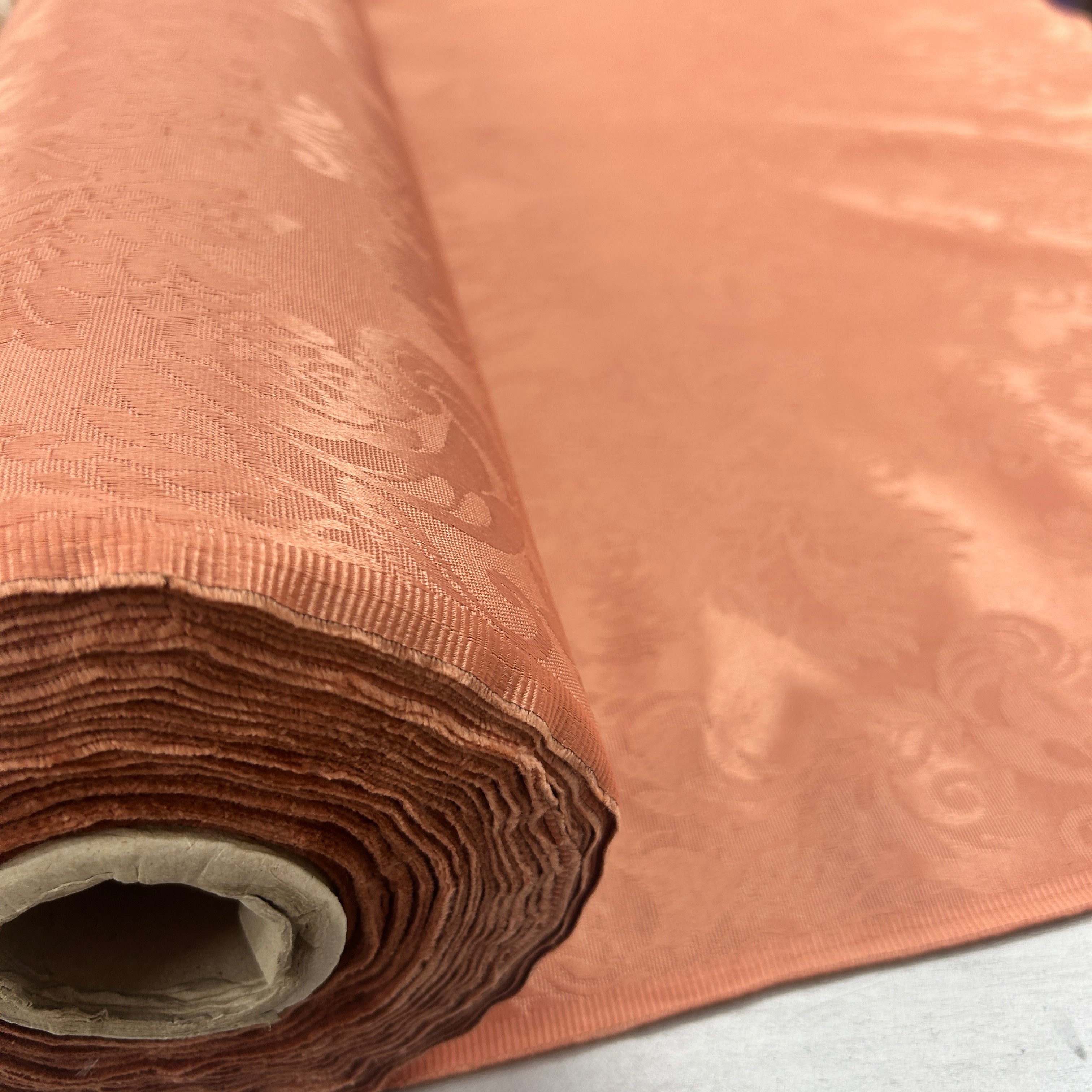 A close-up of a rolled peach-colored damask fabric with a subtle woven pattern, showing its texture and color variation.