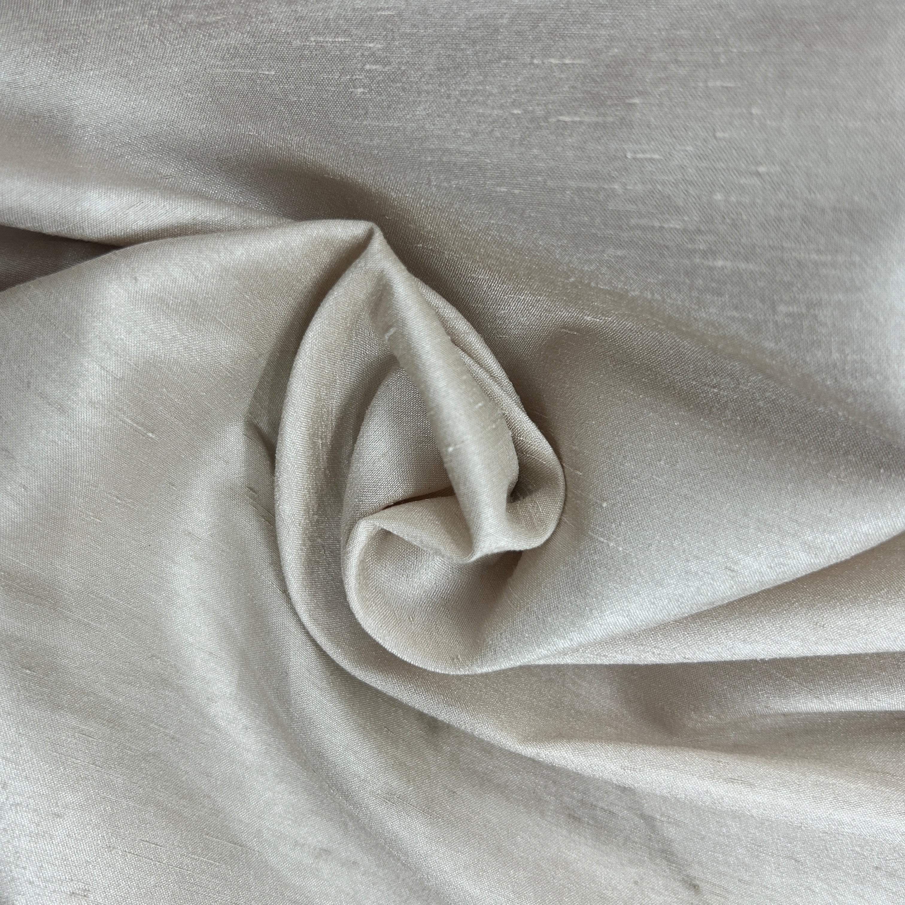 A close-up of draped light beige fabric with a soft, slightly lustrous texture and gentle folds.