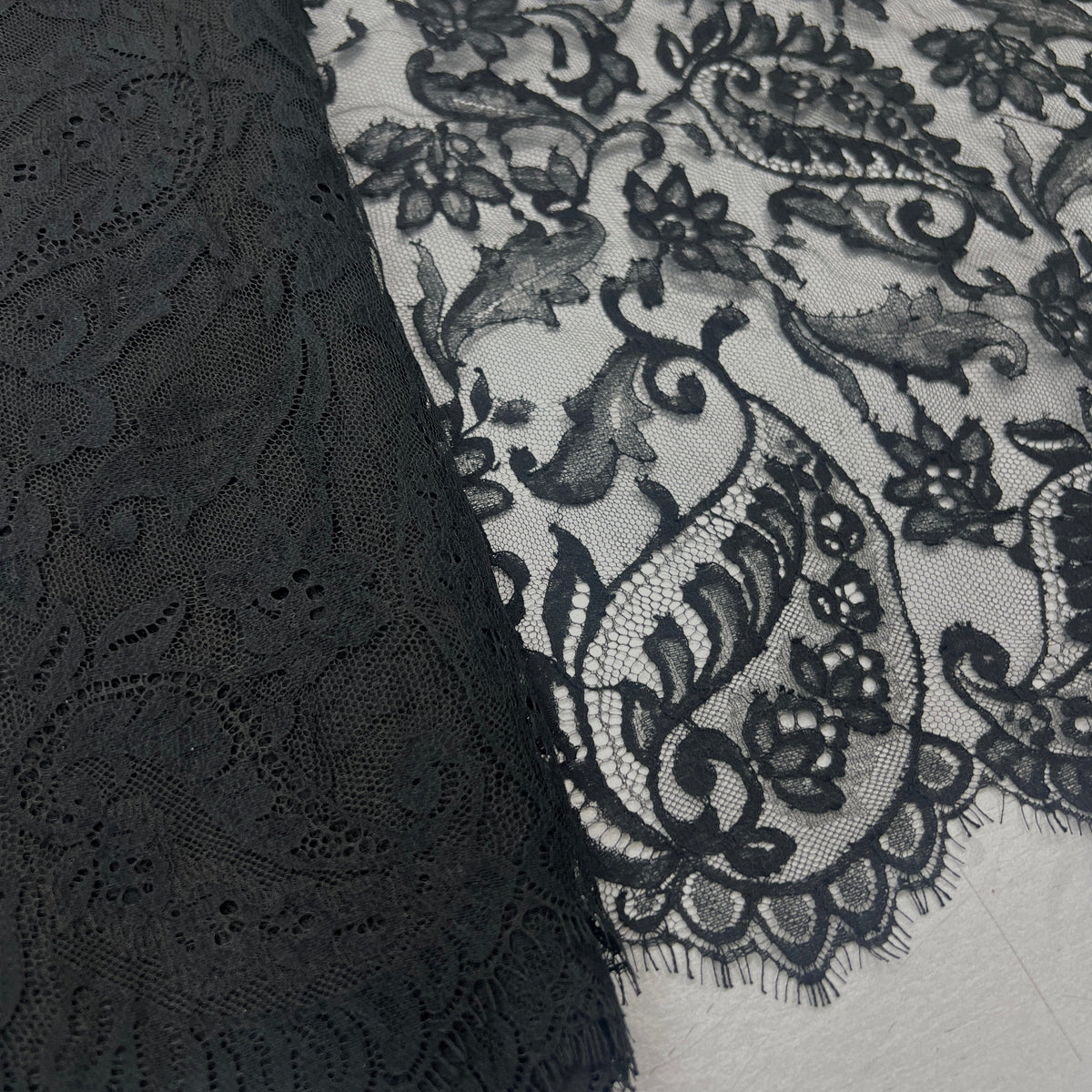 Black lace fabric with an intricate floral and paisley pattern, shown draped over a white surface.