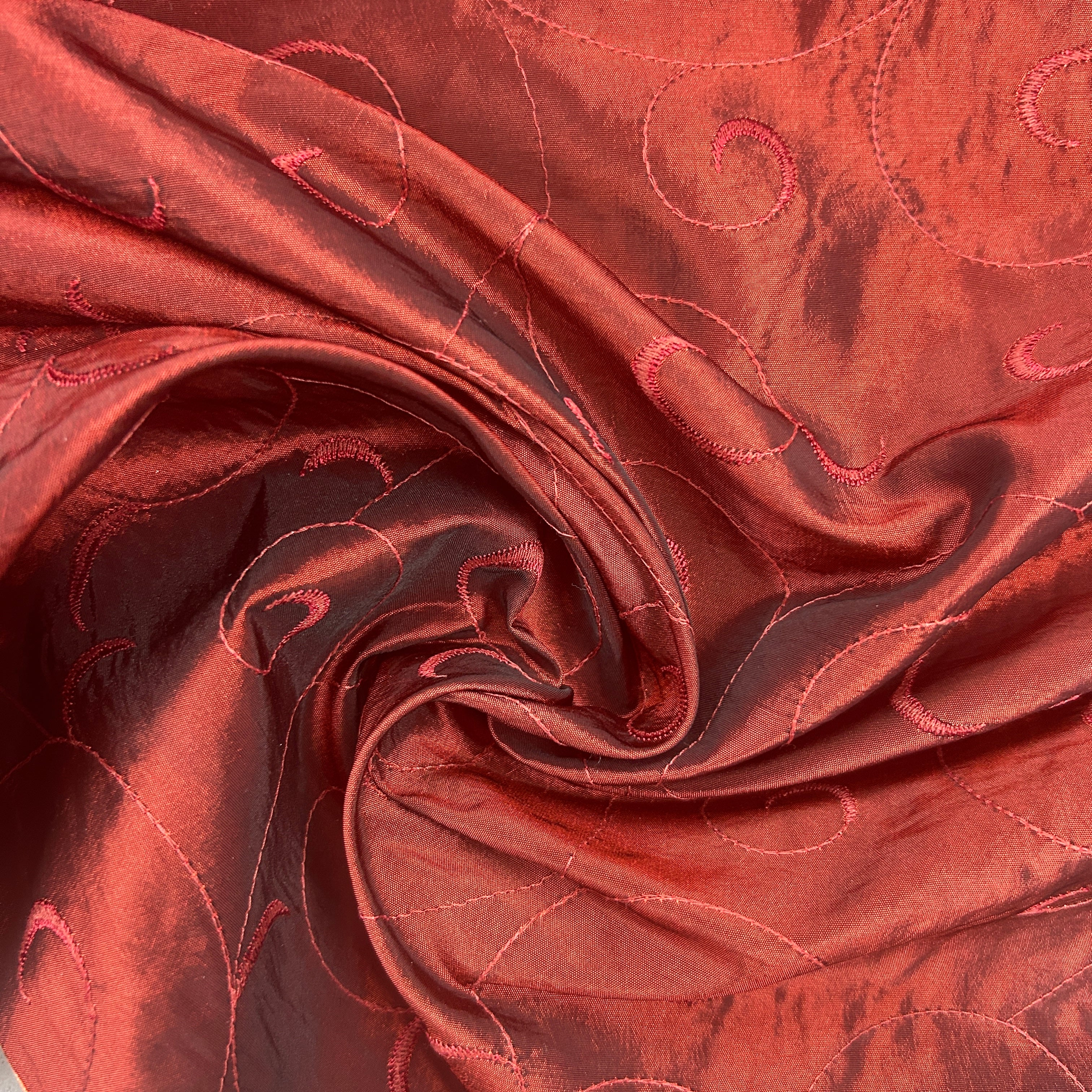 A close-up of draped red fabric with a subtle sheen and stitched swirl pattern, arranged in a spiral fold.