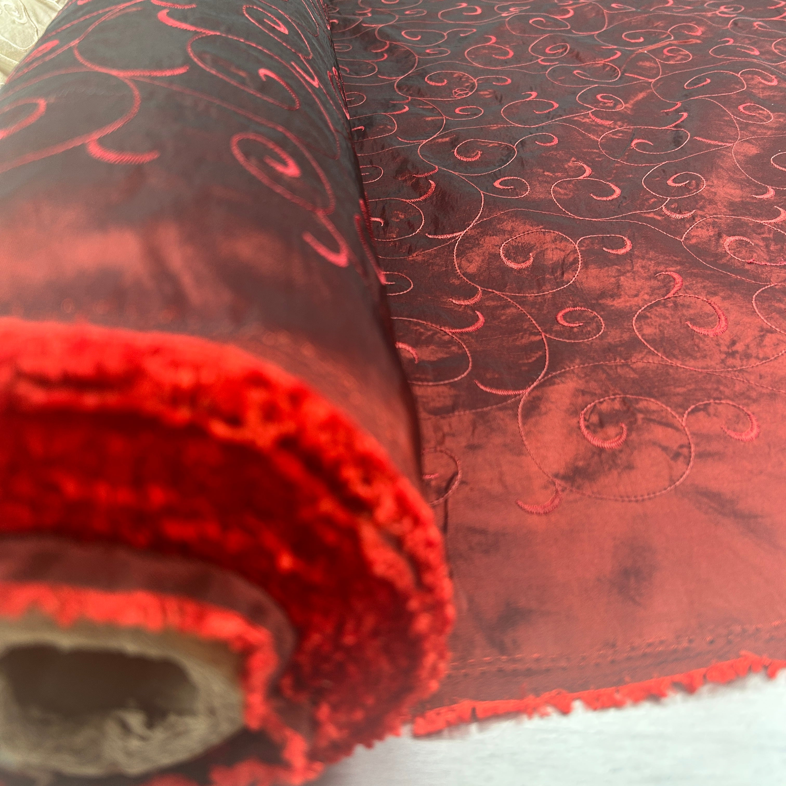 A close-up of a rolled piece of red velvet fabric with a tonal embroidered swirl pattern, showing the plush texture and the inner backing.