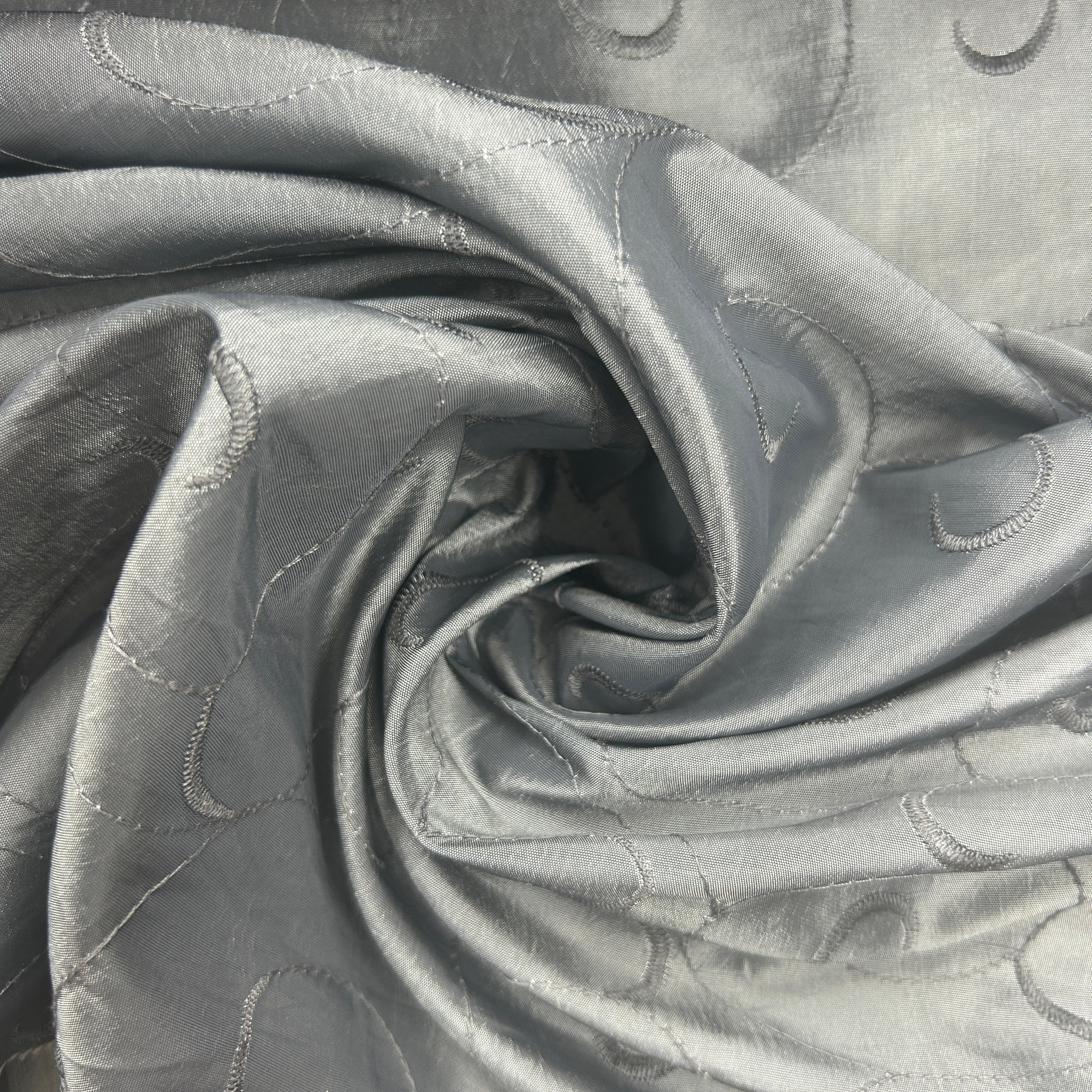 A close-up of a light gray fabric with a subtle sheen, featuring stitched wave-like patterns and draped in a spiral arrangement.