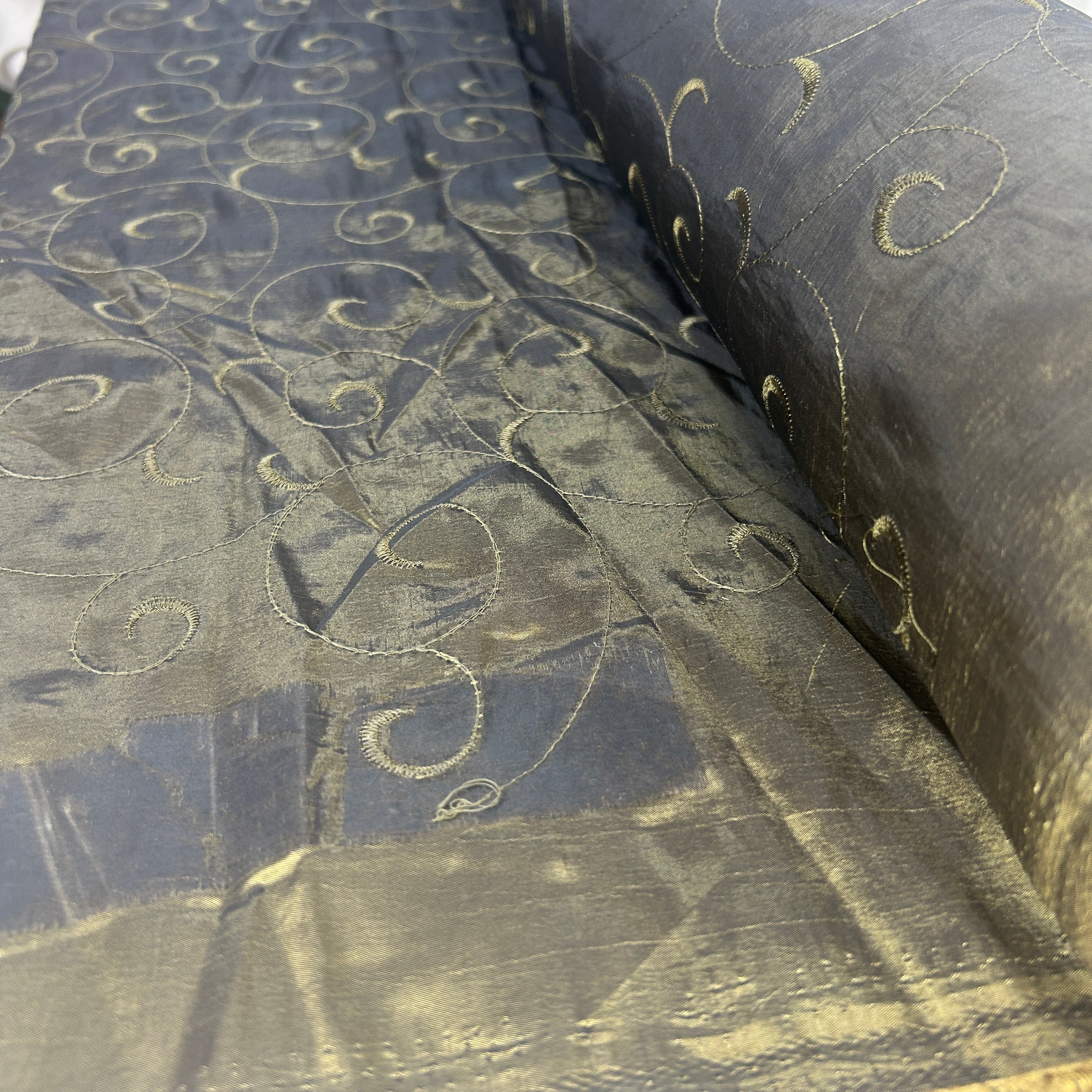 A close-up of a sheer black fabric with a shimmering gold metallic swirl pattern, draped to show its texture and sheen.