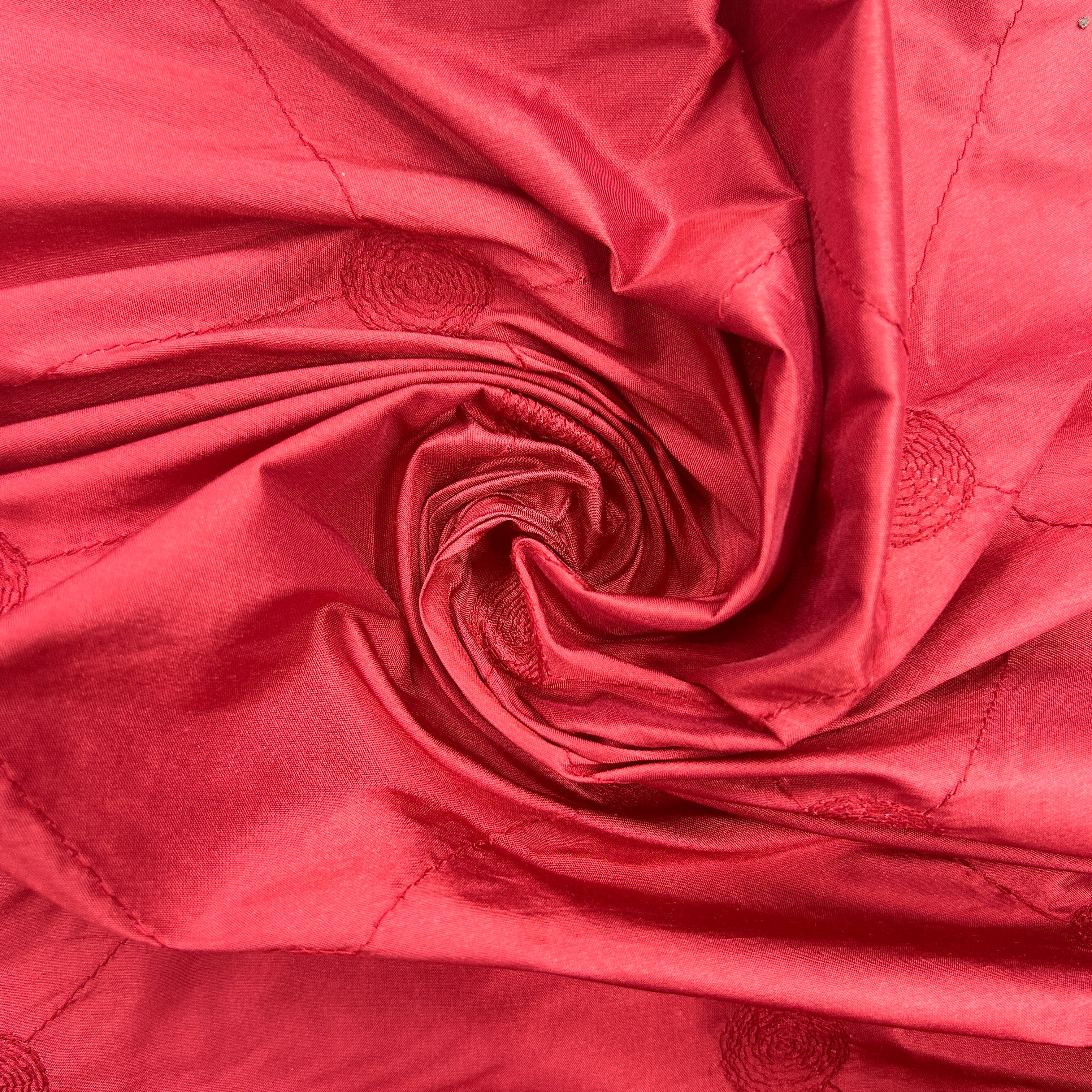 A close-up of draped red fabric with a spiral twist in the center and subtle tonal circular embroidered motifs.