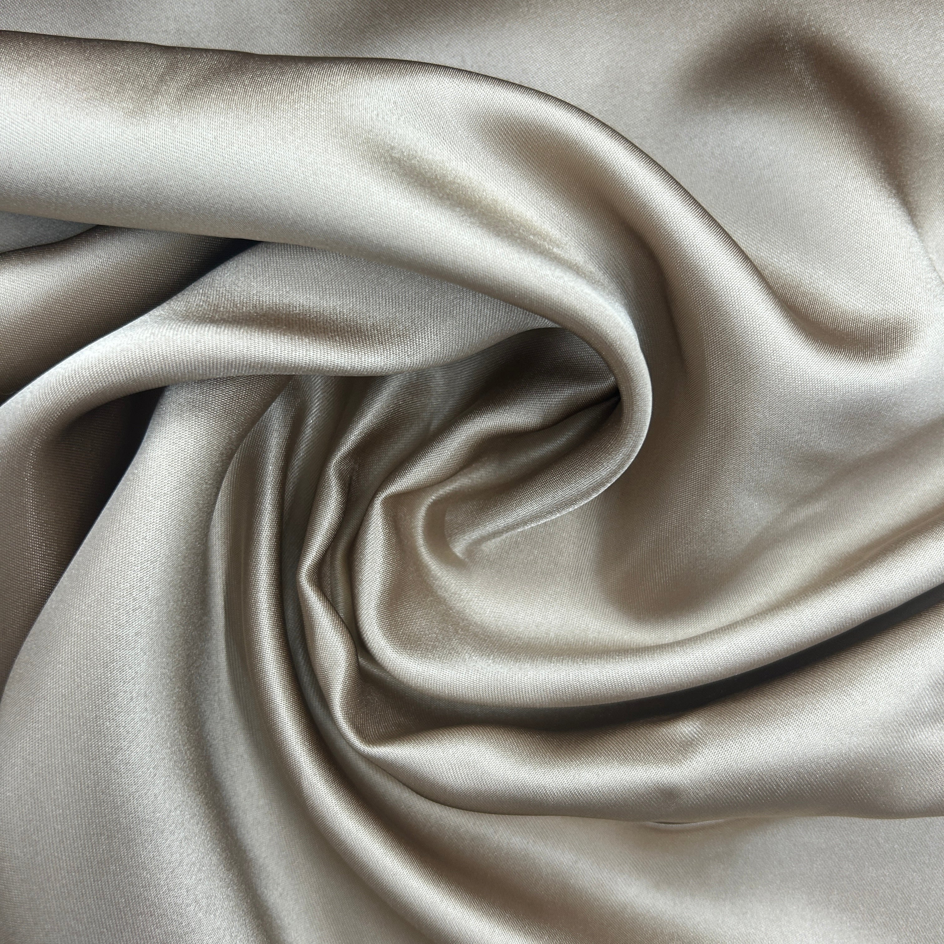 A close-up of draped satin fabric in a warm taupe-beige color, showing its smooth texture and soft sheen.
