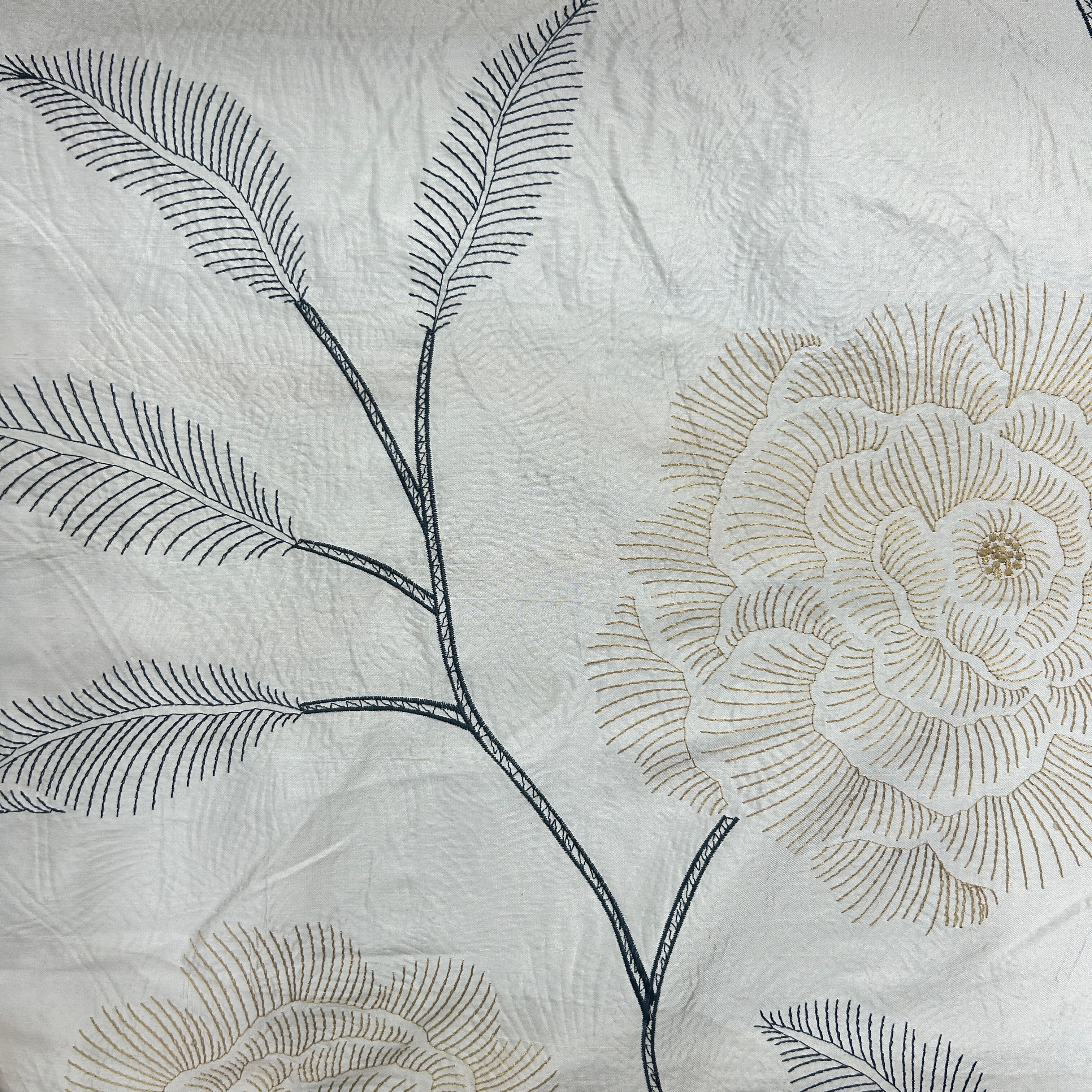 A close-up of a white fabric surface with embroidered floral and leaf motifs in dark blue-gray and warm beige/tan thread.