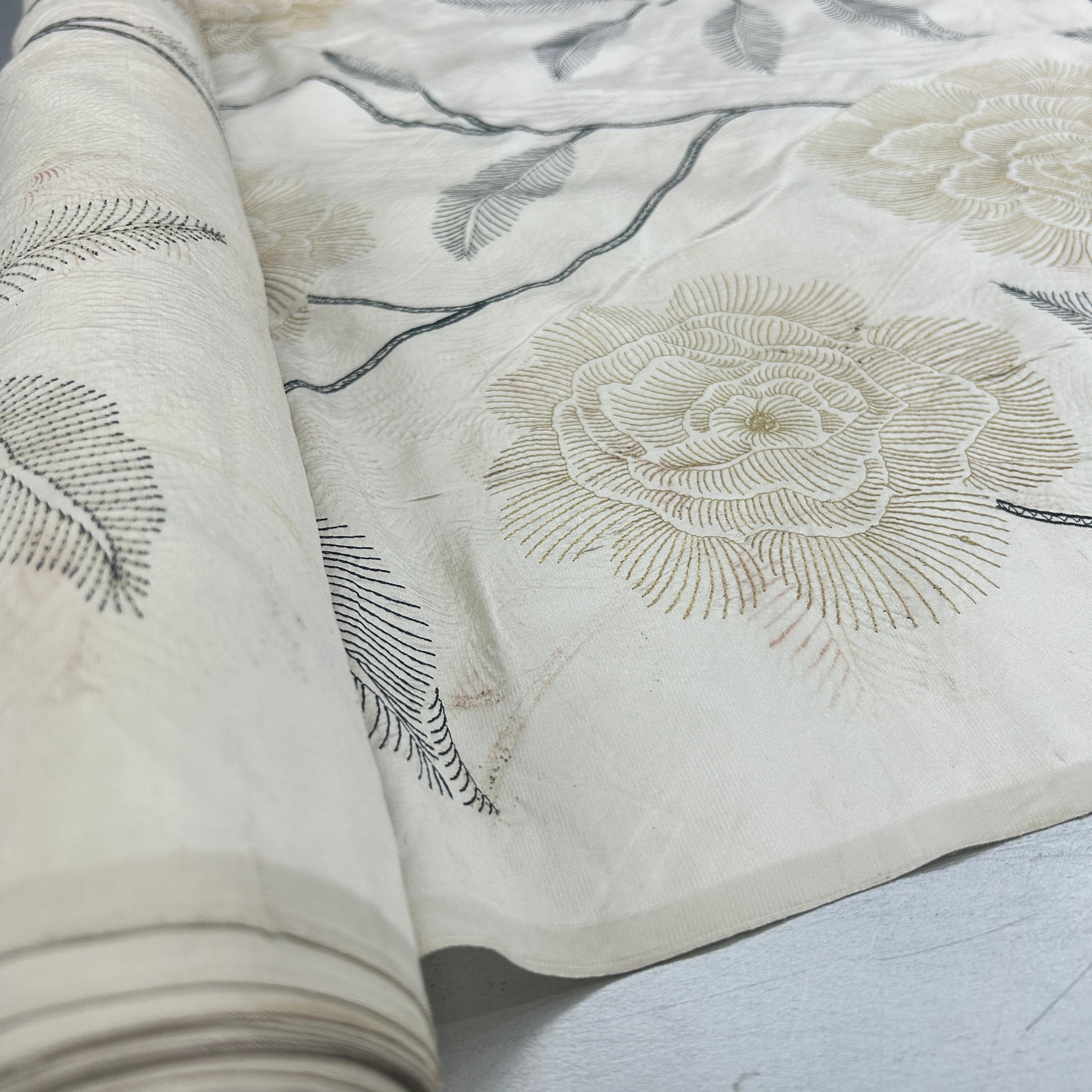 A close-up of a rolled piece of cream-colored fabric with embroidered floral and leaf patterns in beige and dark gray thread, laid over a light gray surface.