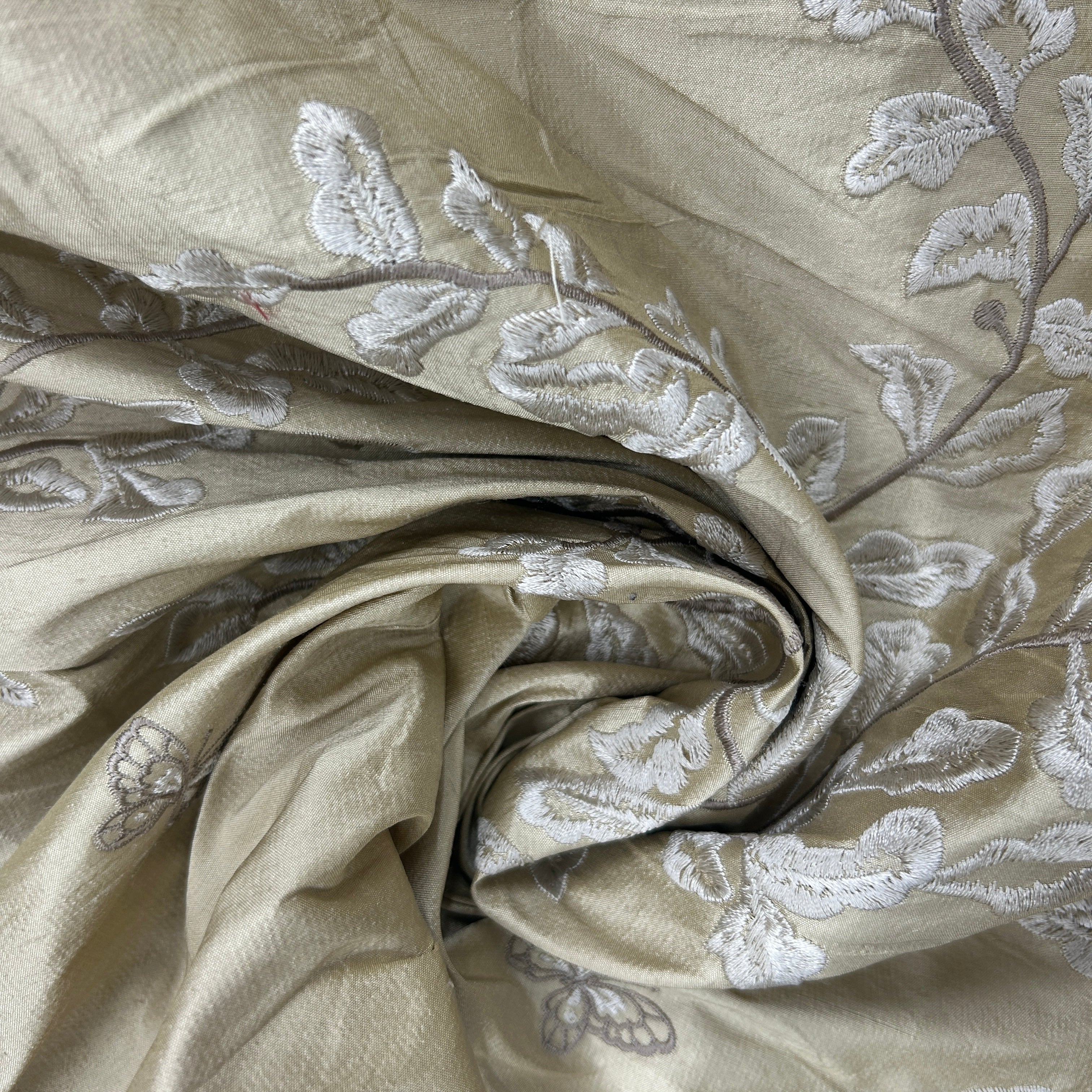 A close-up of draped fabric in a warm beige or champagne tone with embroidered floral and butterfly motifs in light gray and white.