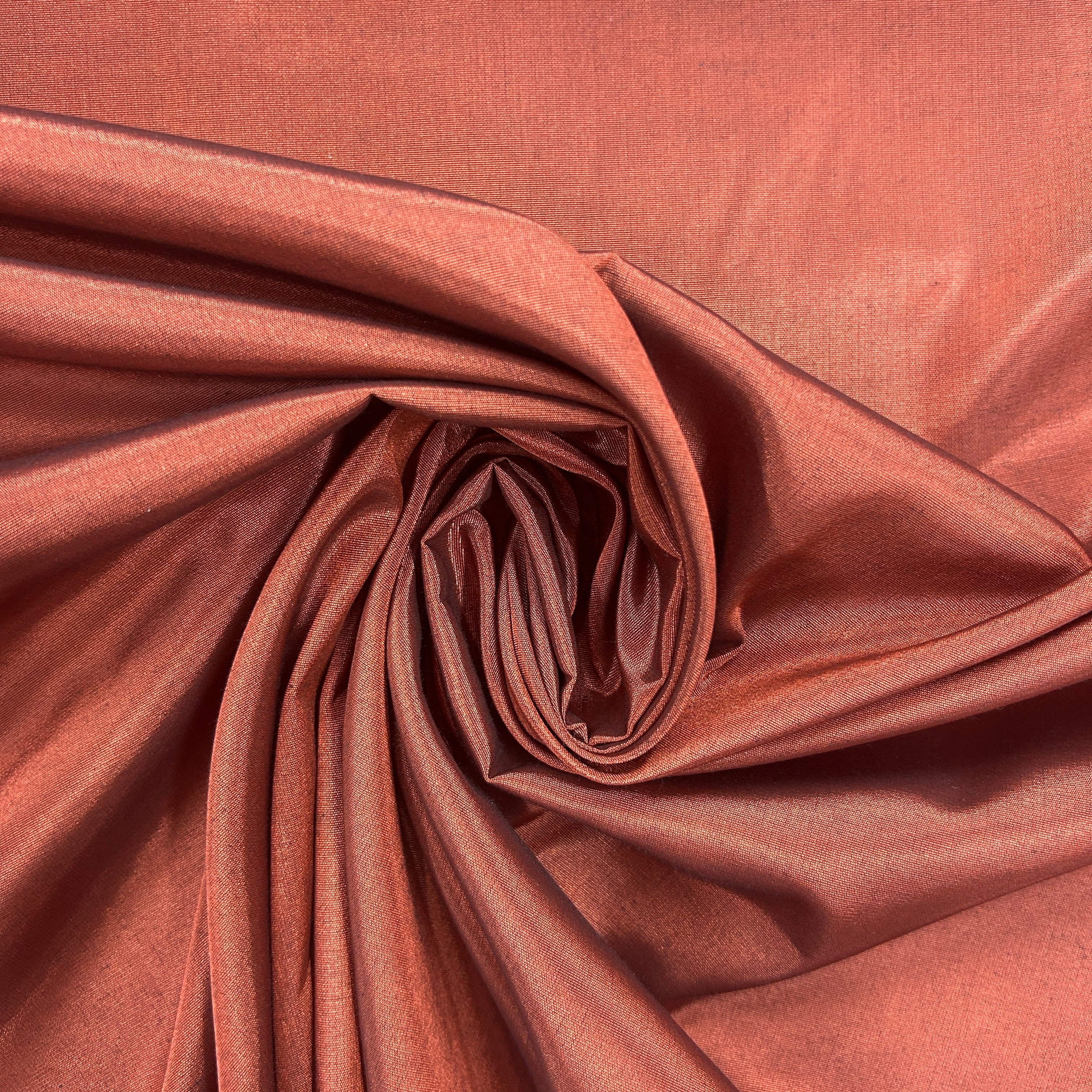 A close-up of draped fabric in a warm coppery rose tone, arranged in a spiral fold pattern that highlights its smooth texture and sheen.