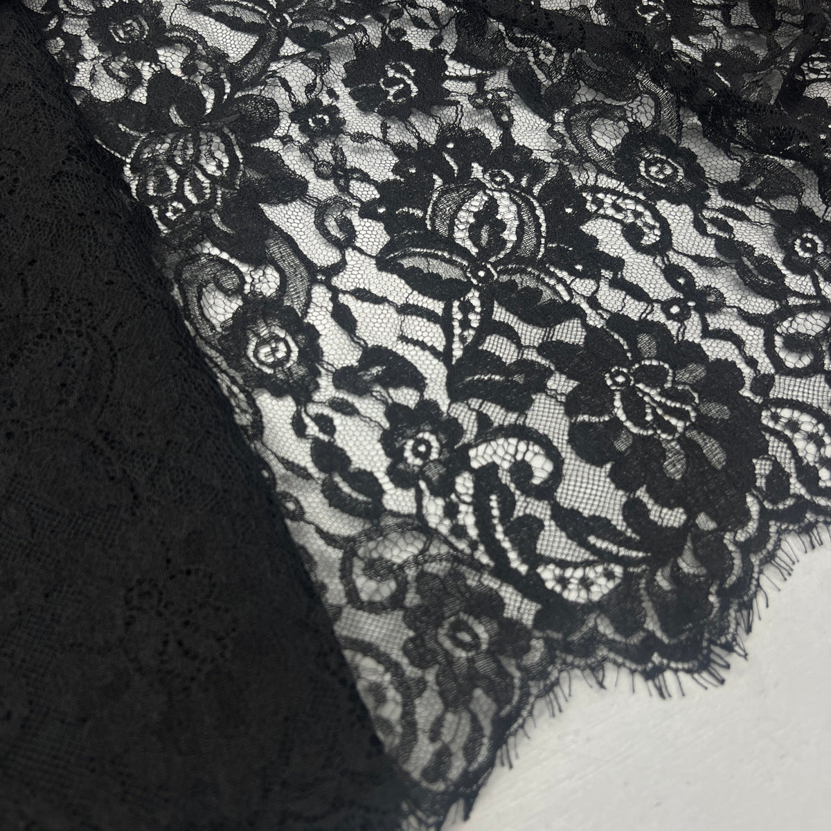 Black floral lace fabric with a sheer mesh background and frayed edges, shown folded on a light surface.