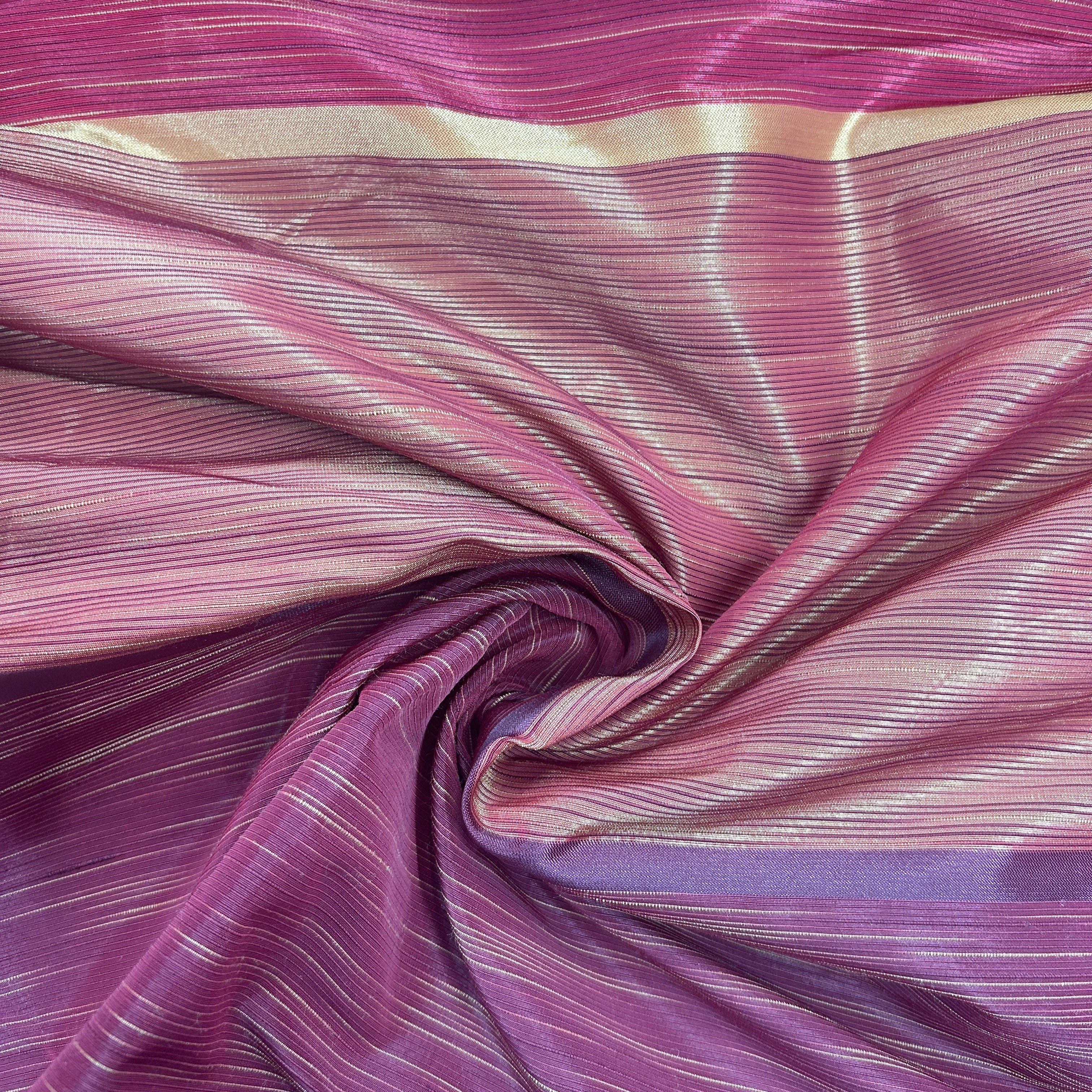 A close-up of a draped fabric with a shimmering magenta and purple colorway, featuring fine horizontal metallic gold stripes and a central swirl.