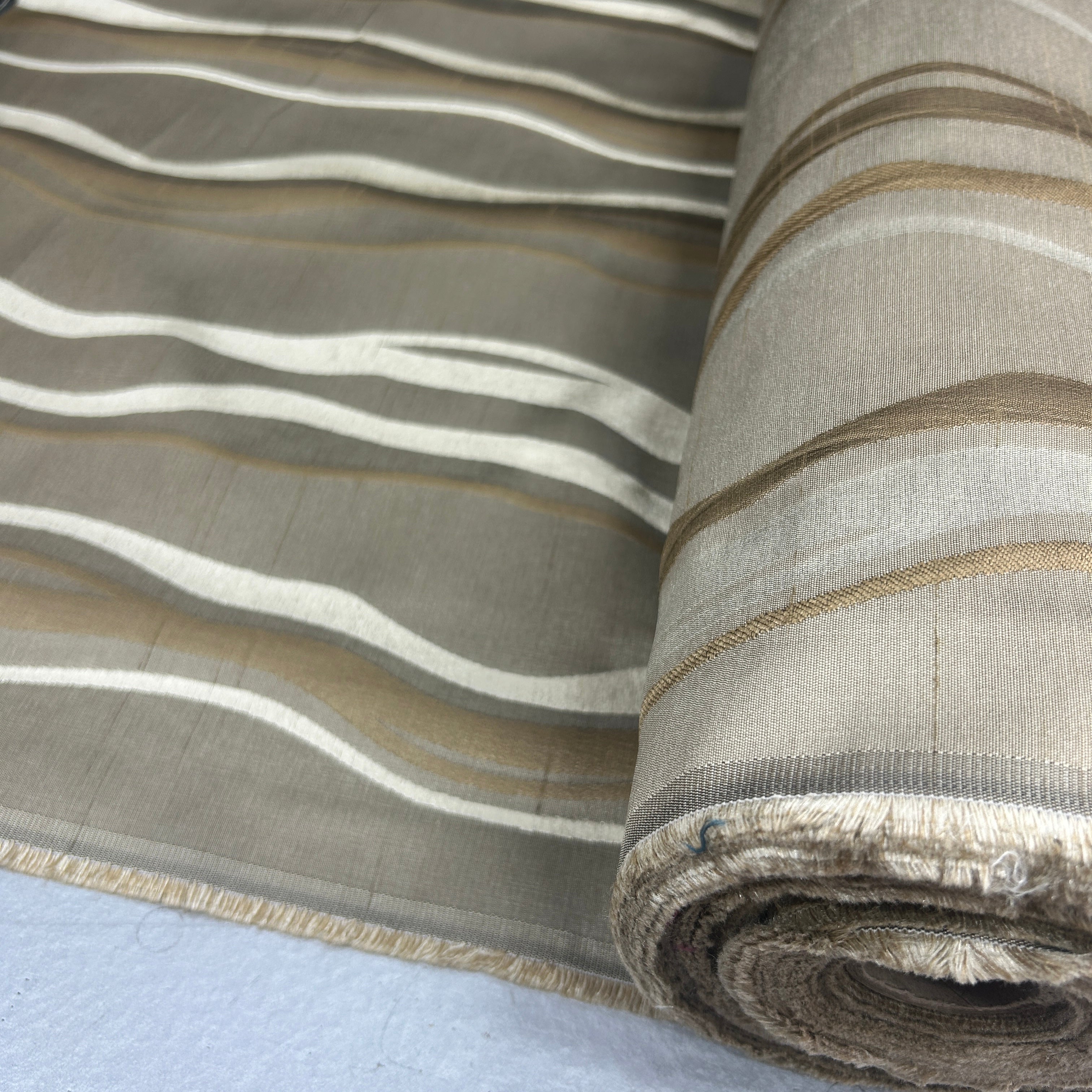 A close-up of a rolled piece of woven fabric with wavy horizontal stripes in taupe, beige, and off-white, showing the textured weave and frayed edge.