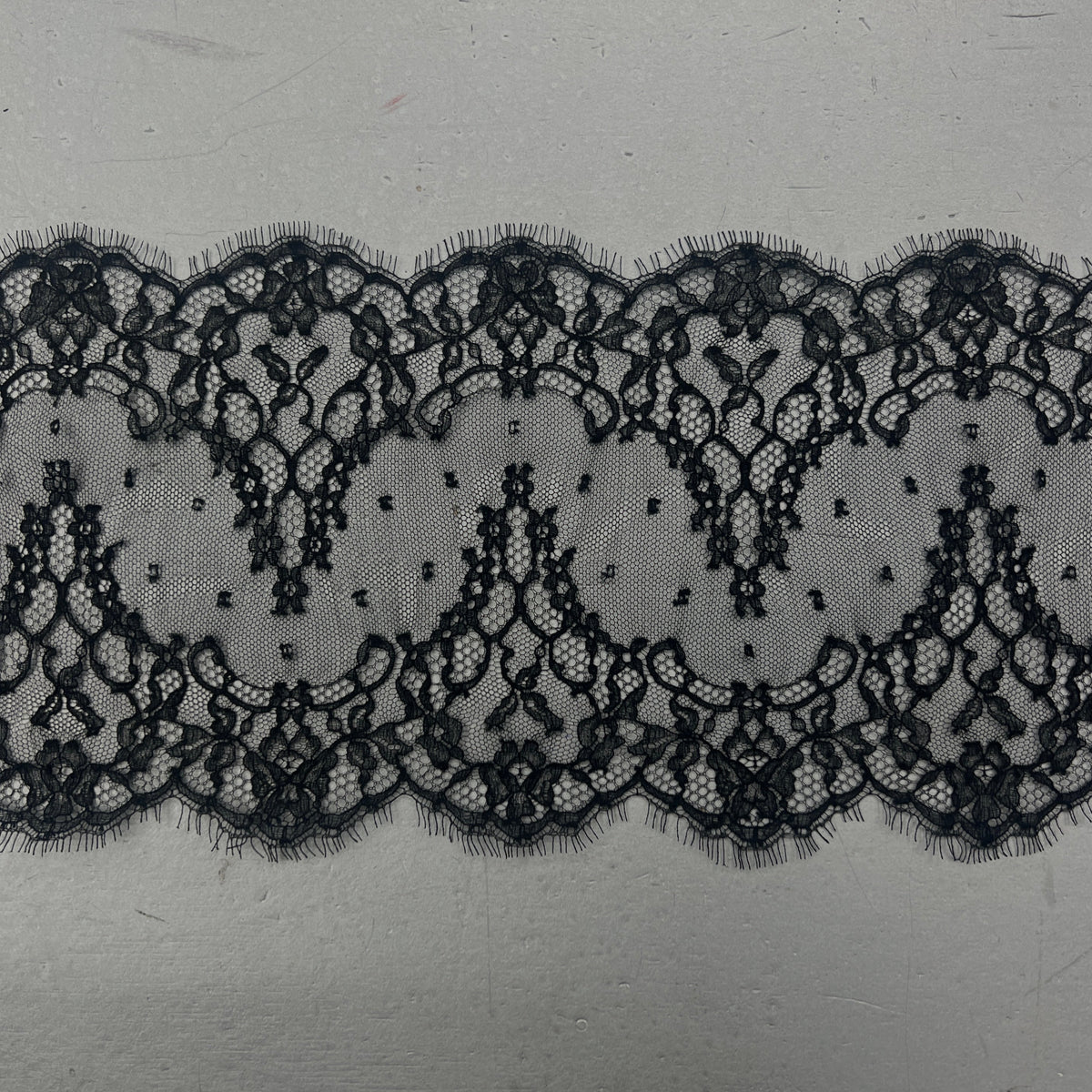 A close-up of a black lace trim with an intricate floral pattern and scalloped edges, laid horizontally on a light gray textured surface.