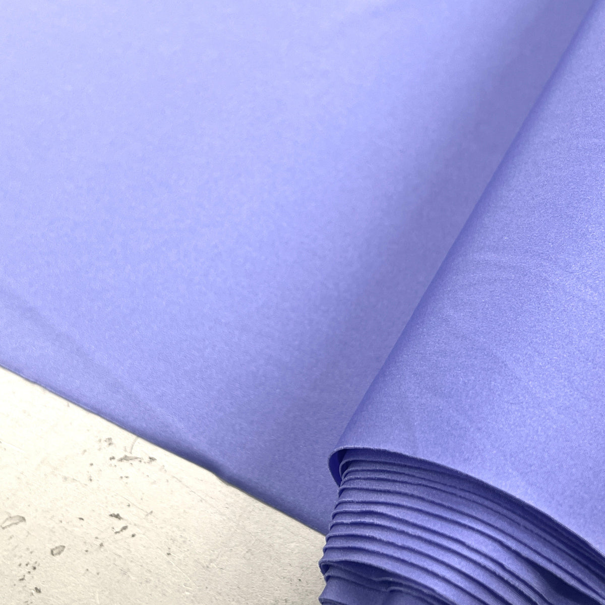 A close-up of light periwinkle blue fabric, partially rolled, resting on a light gray concrete surface.
