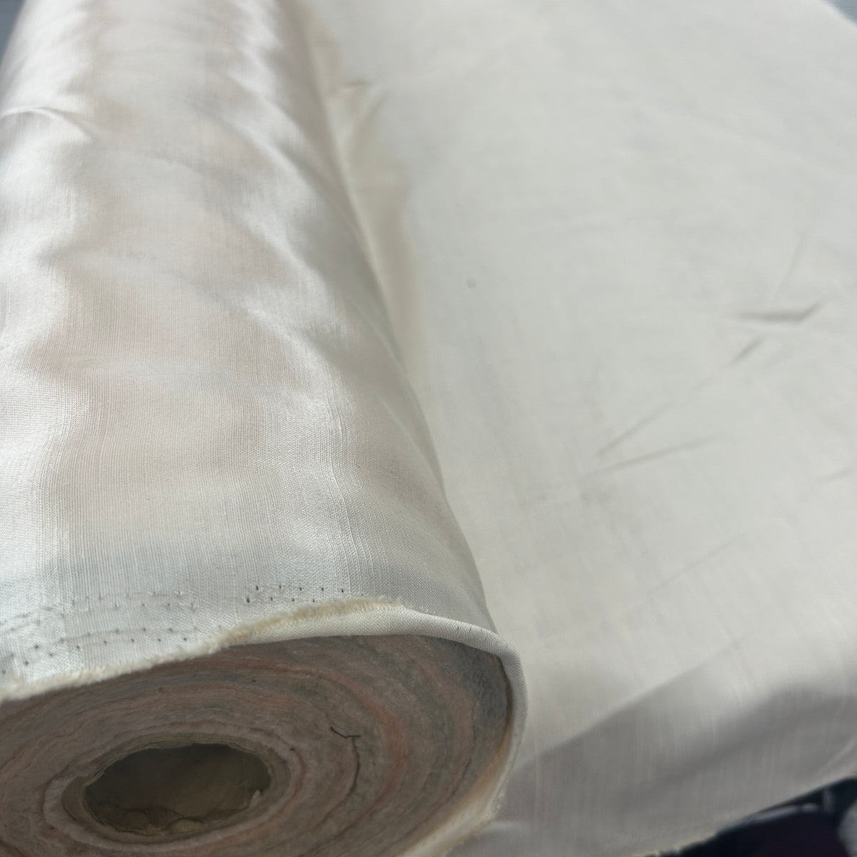 A close-up of a rolled white fabric or material, showing its smooth texture and slight sheen, with the unrolled section laid out behind it.