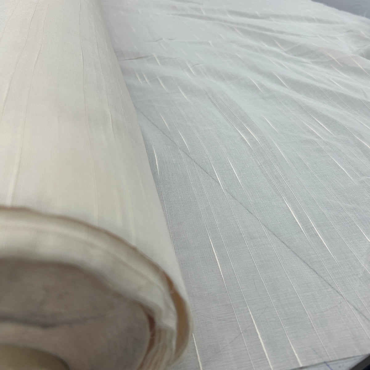A close-up of a rolled off-white fabric or textile with a subtle woven texture and fine vertical striations, shown against a matching light background.