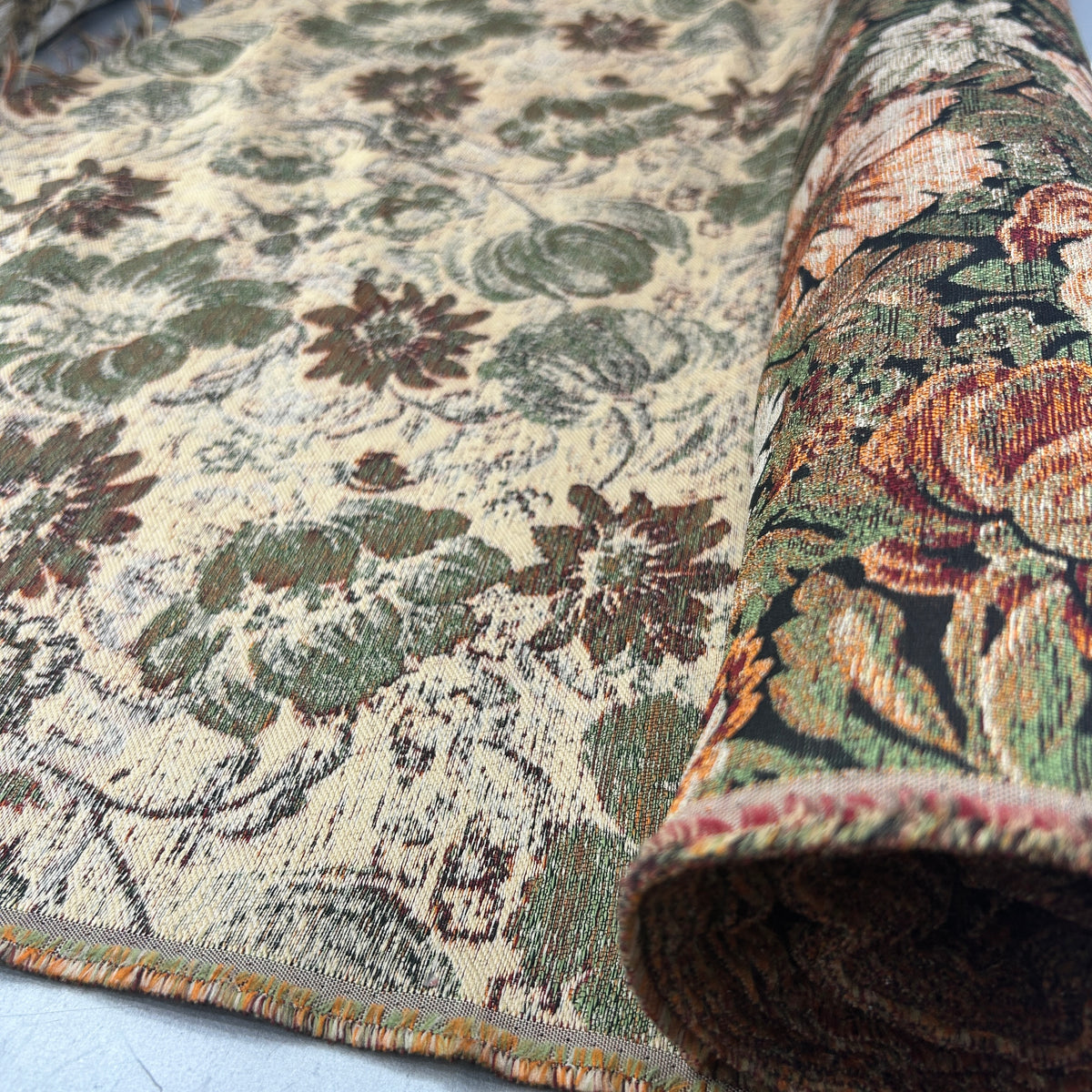 A close-up of a patterned area rug with one end rolled back to show the underside and pile. The rug features a vintage floral design in muted tones of green, brown, and rust orange on a warm beige background.