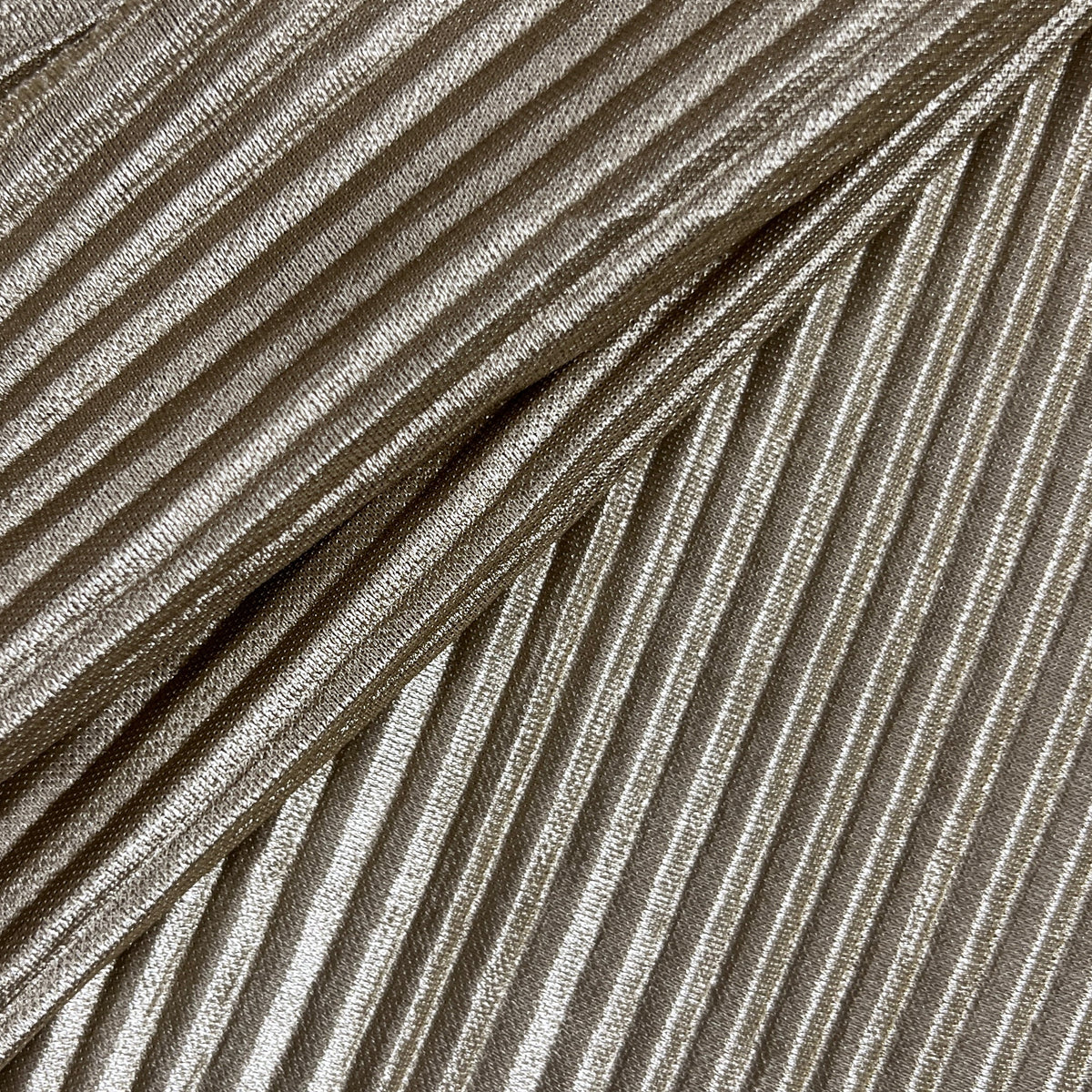 A close-up of a pleated fabric or material sample with narrow vertical pleats in a warm taupe-beige tone, photographed against a light neutral background.