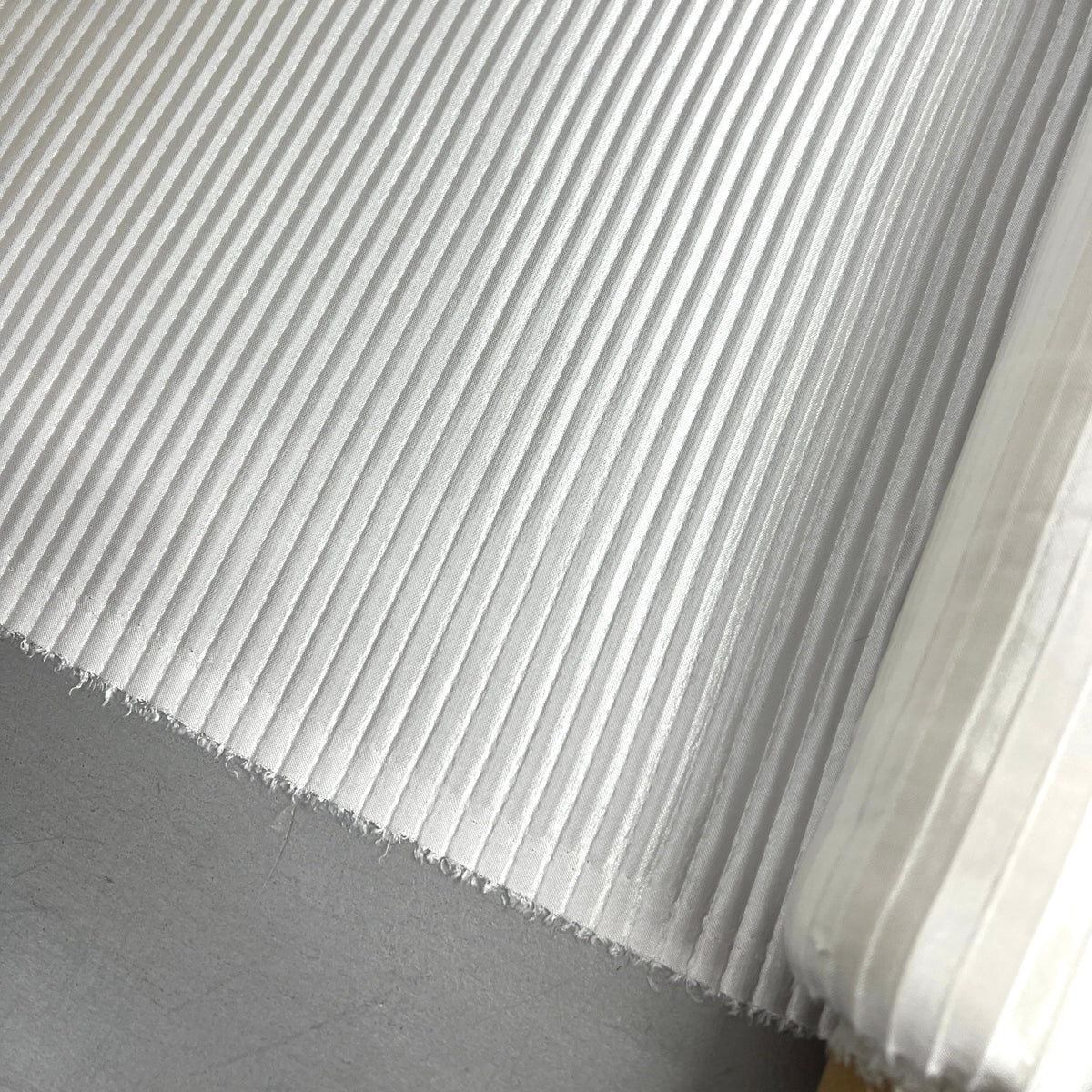 A close-up of a white pleated or ribbed fabric/material surface with a frayed edge, photographed against a gray floor background.