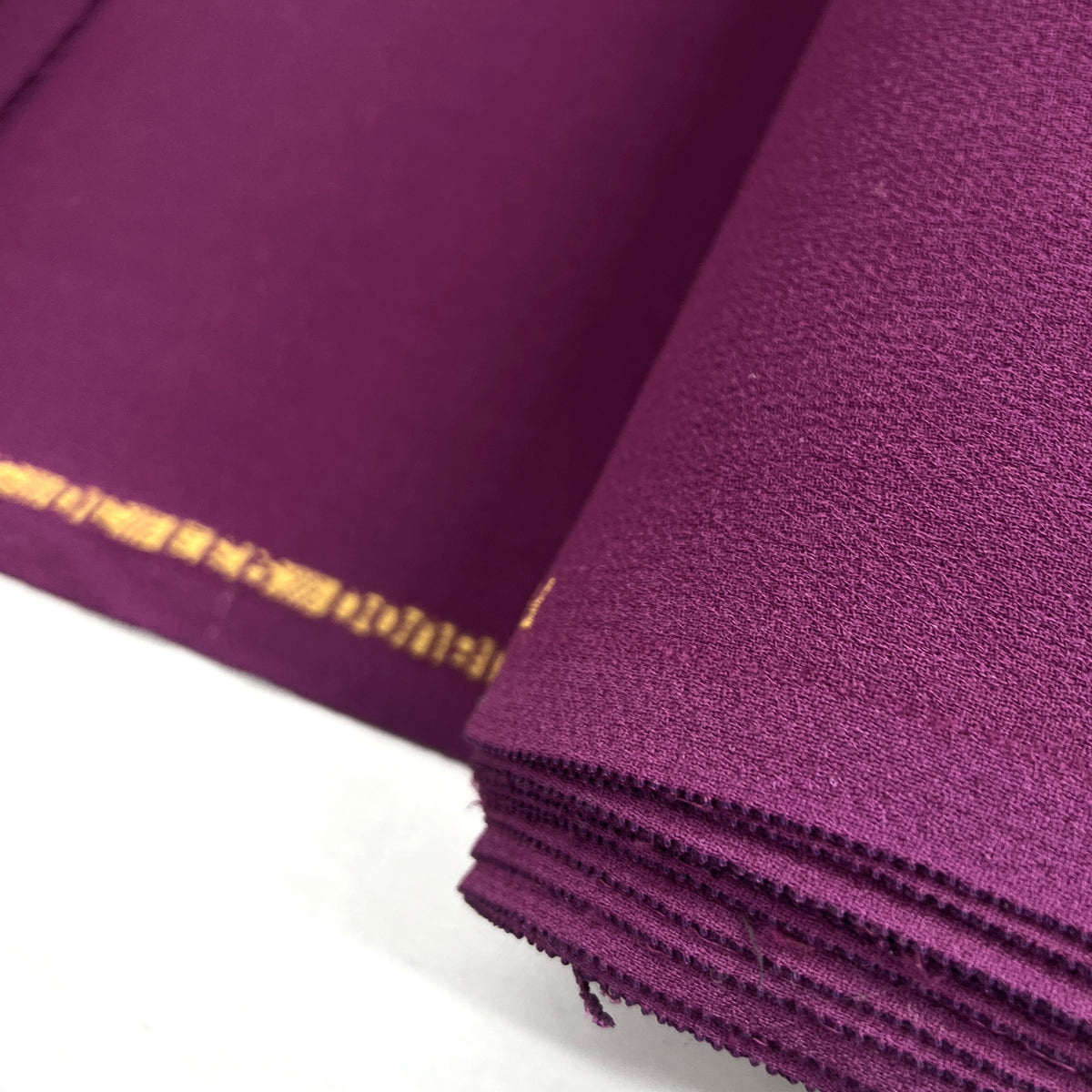 A close-up of folded purple fabric with a visible gold border along one edge, photographed on a white surface.