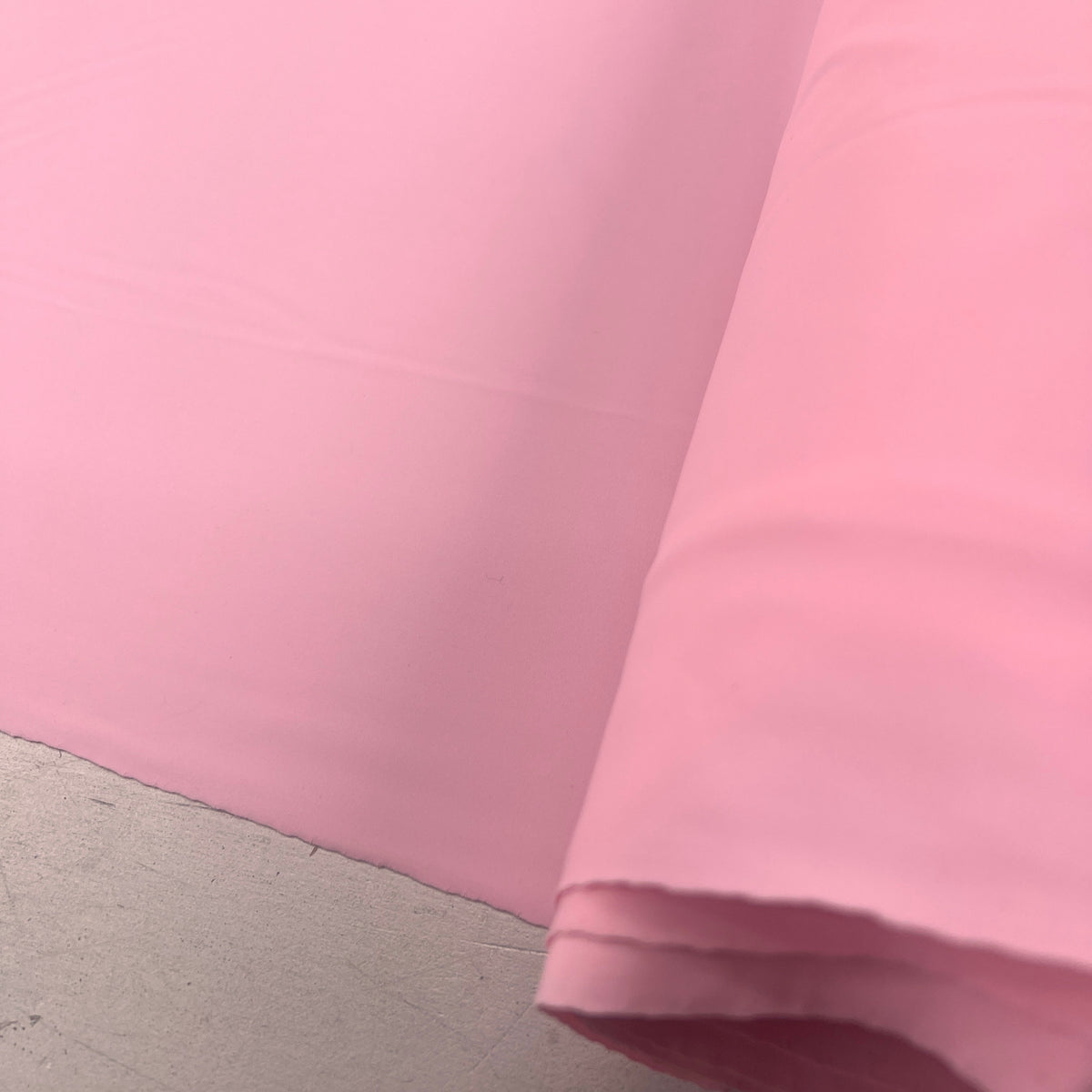 A close-up of a roll of solid light pink fabric, showing its smooth texture and drape against a neutral gray concrete floor.