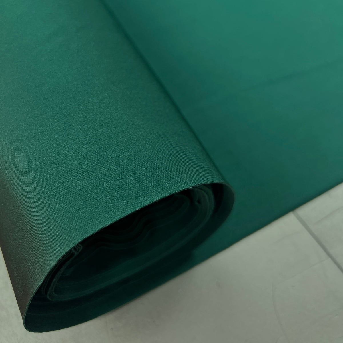 A roll of dark teal green fabric or material, partially unrolled to show its texture, set against a light gray tiled floor.
