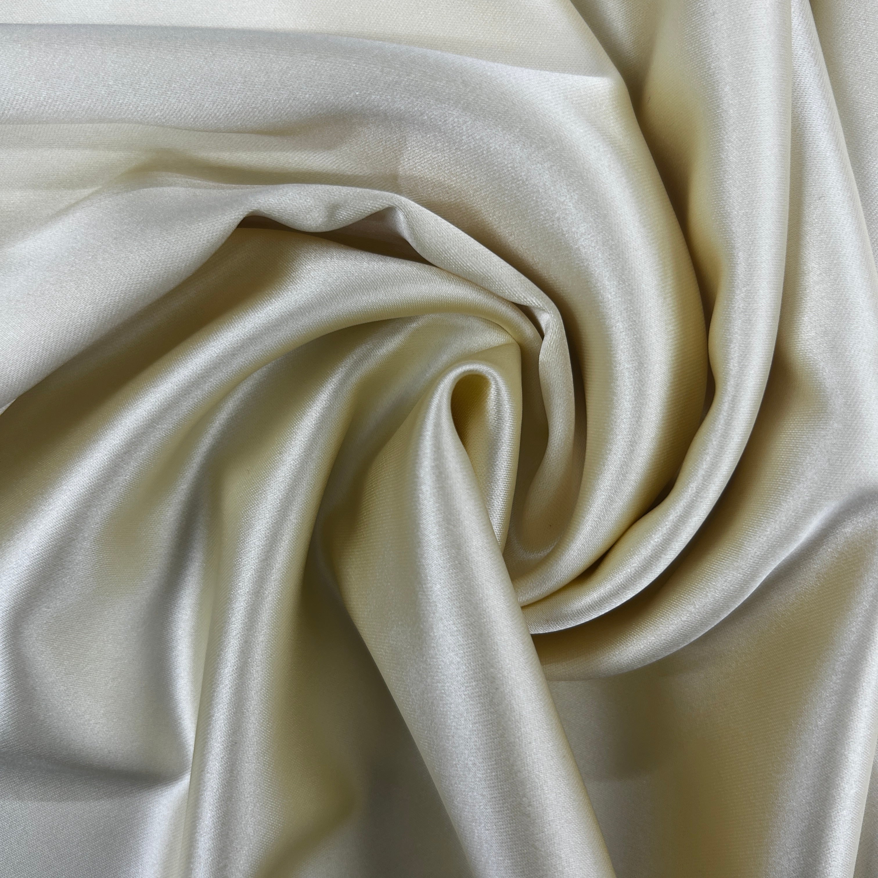 A close-up of draped satin fabric in a warm ivory or cream color, arranged in a spiral pattern to show off its smooth, lustrous texture.