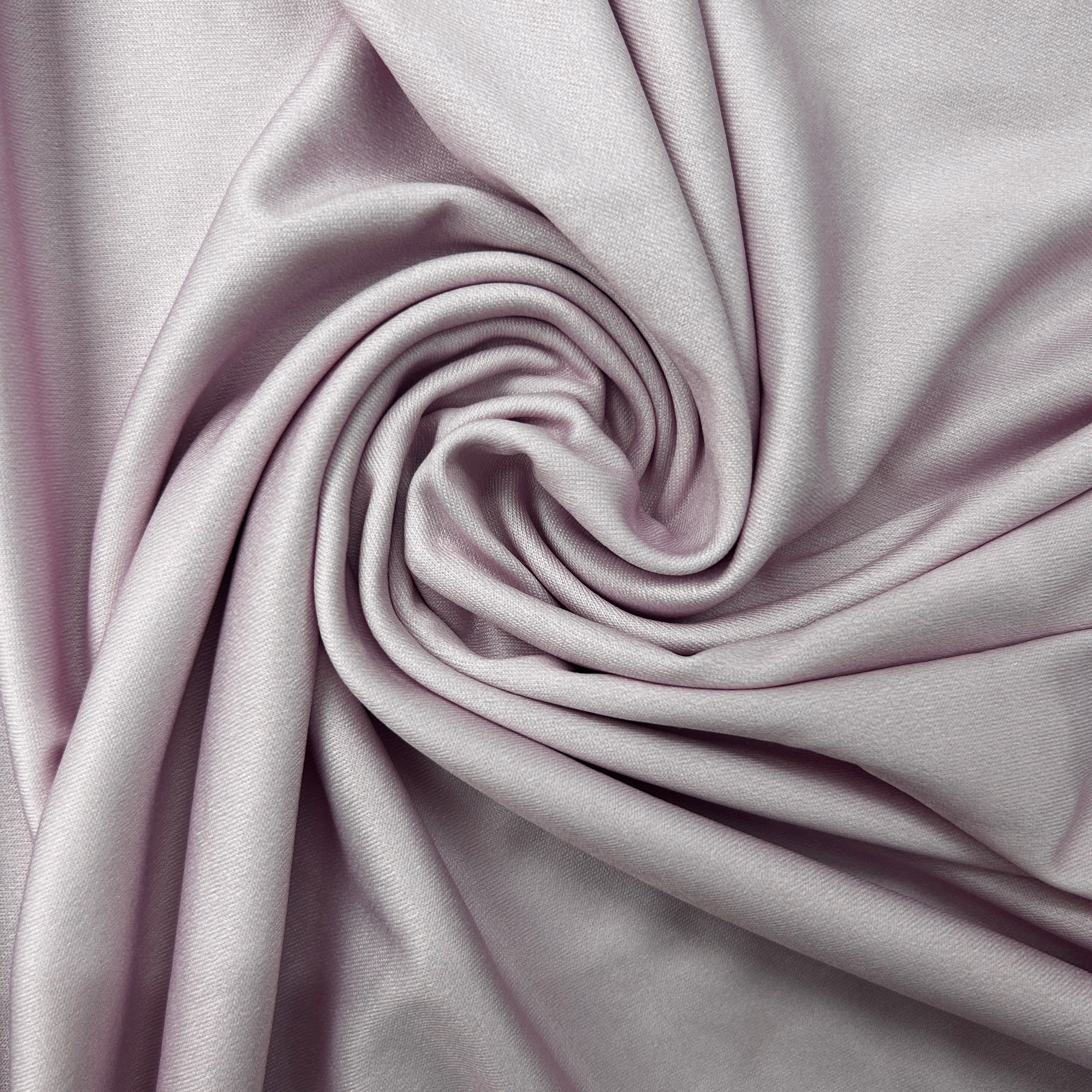 A close-up of draped satin-like fabric in a soft pale lavender or dusty lilac color, arranged in a spiral fold pattern.