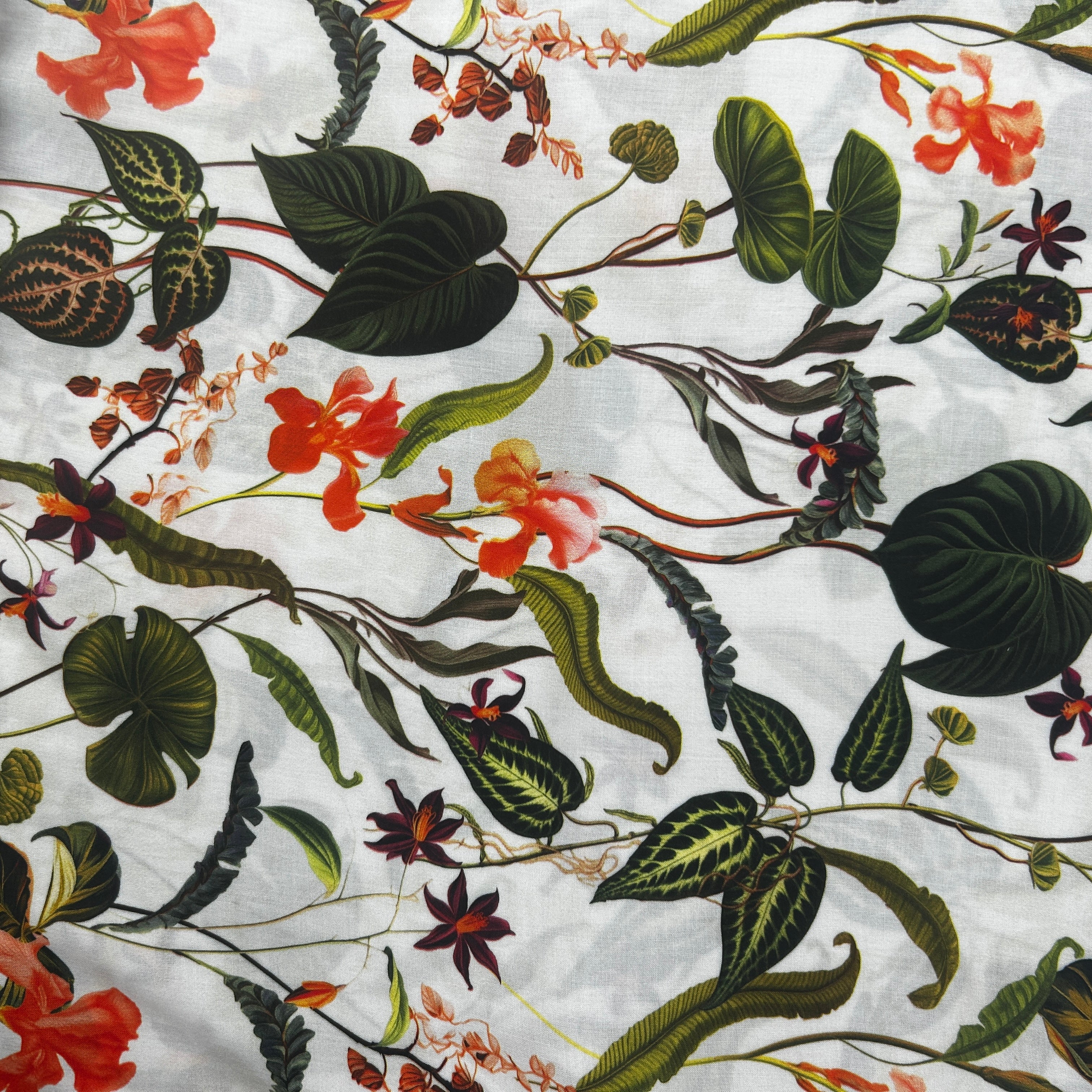 A close-up of a white fabric with an all-over botanical print featuring green leaves, orange flowers, and small dark red accents.