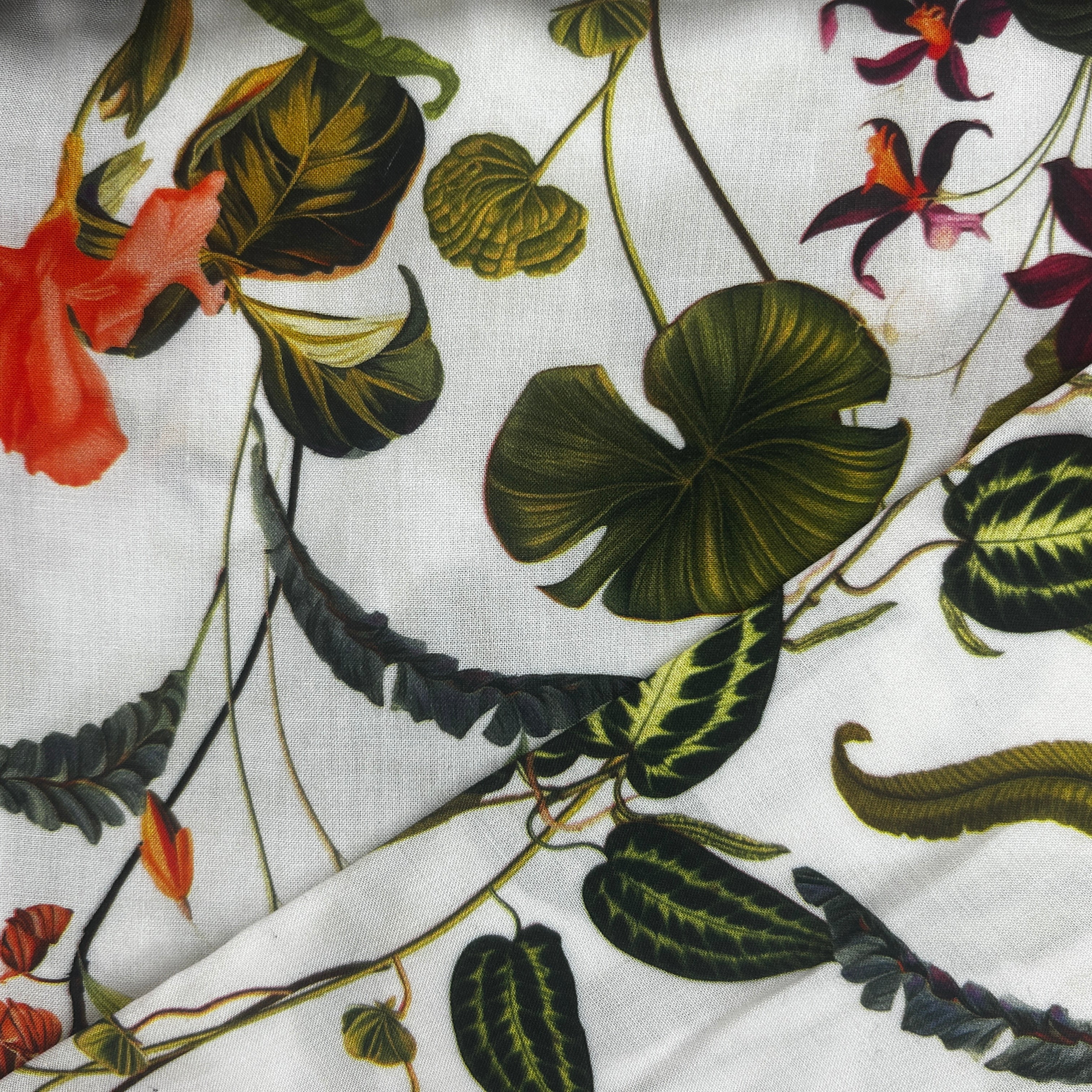 White fabric with a botanical print featuring green monstera and variegated leaves, dark green ferns, and orange and purple flowers.