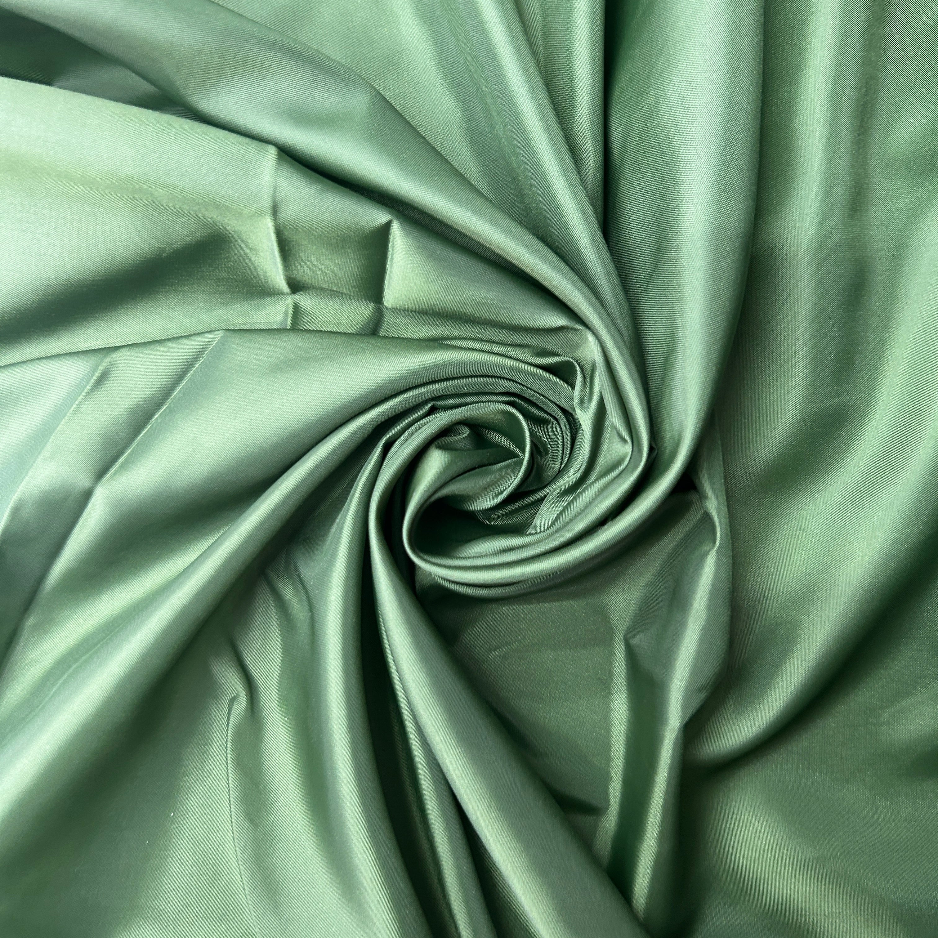 A close-up of draped satin fabric in a muted sage green color, arranged in a spiral pattern to show off its smooth texture and sheen.