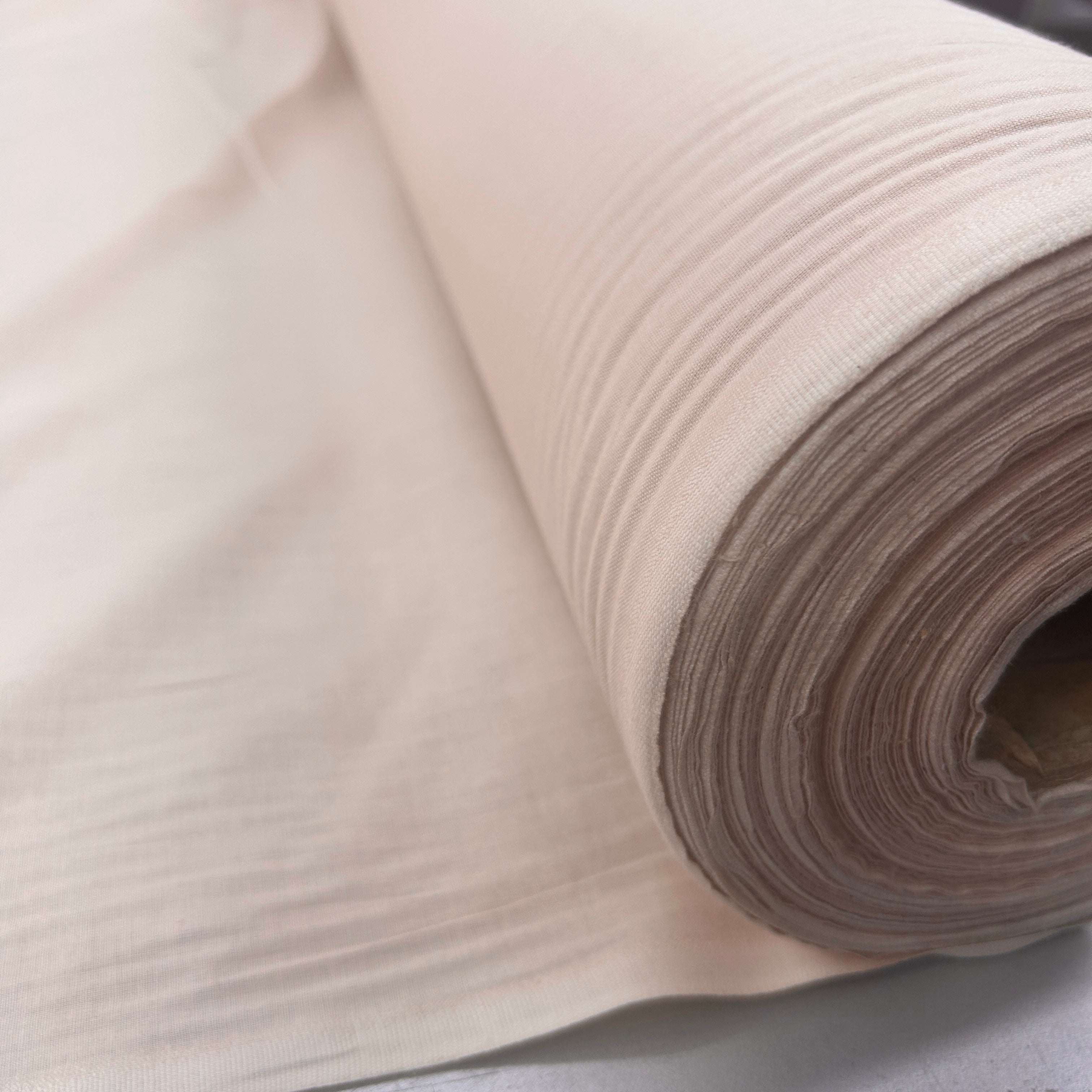 A close-up of a rolled piece of light beige fabric with a subtle horizontal textured weave, shown against a neutral light gray background.