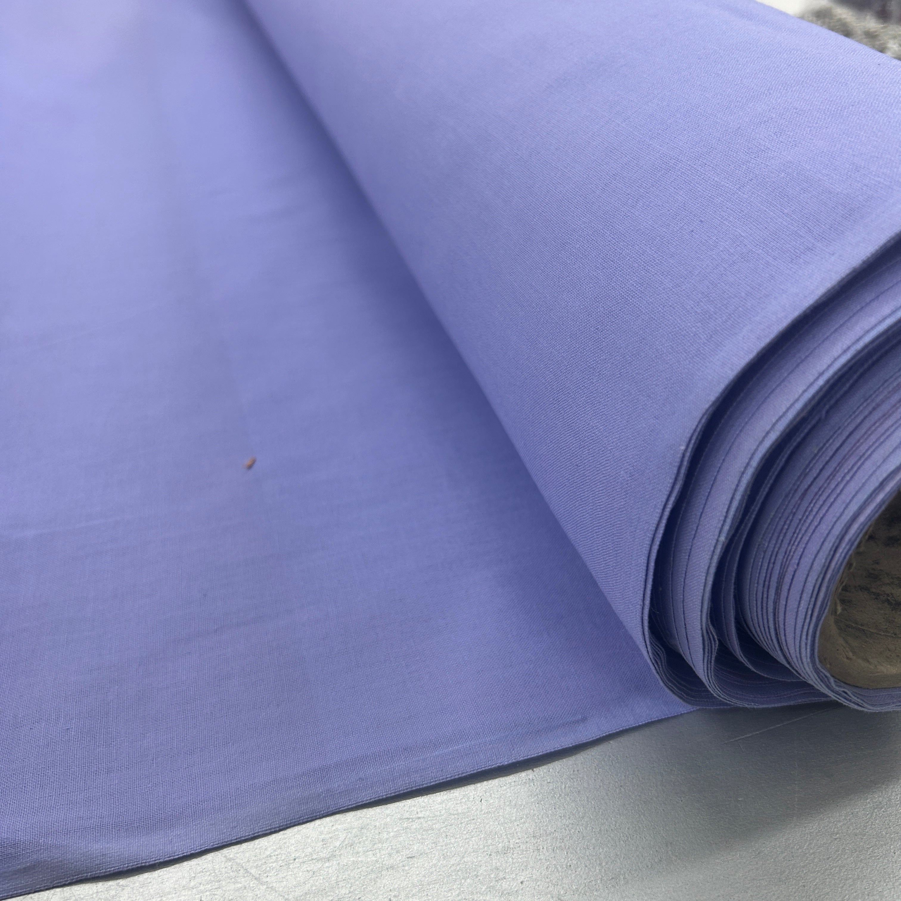 A close-up of a roll of lavender purple fabric, showing its woven texture and drape, resting on a light gray surface.