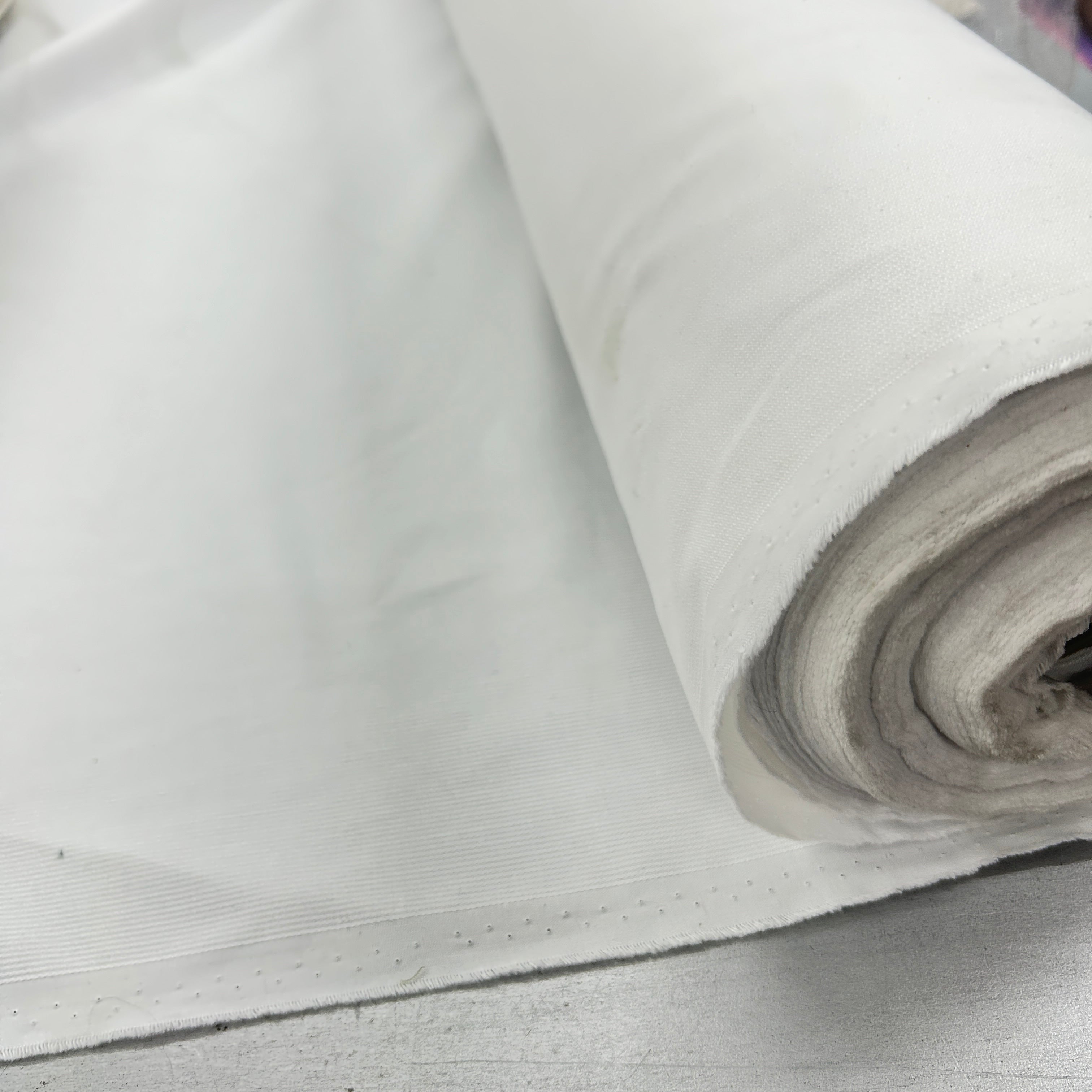 A close-up of a white woven fabric, shown partially unrolled with a rolled section on the right, revealing the texture and weave of the material.