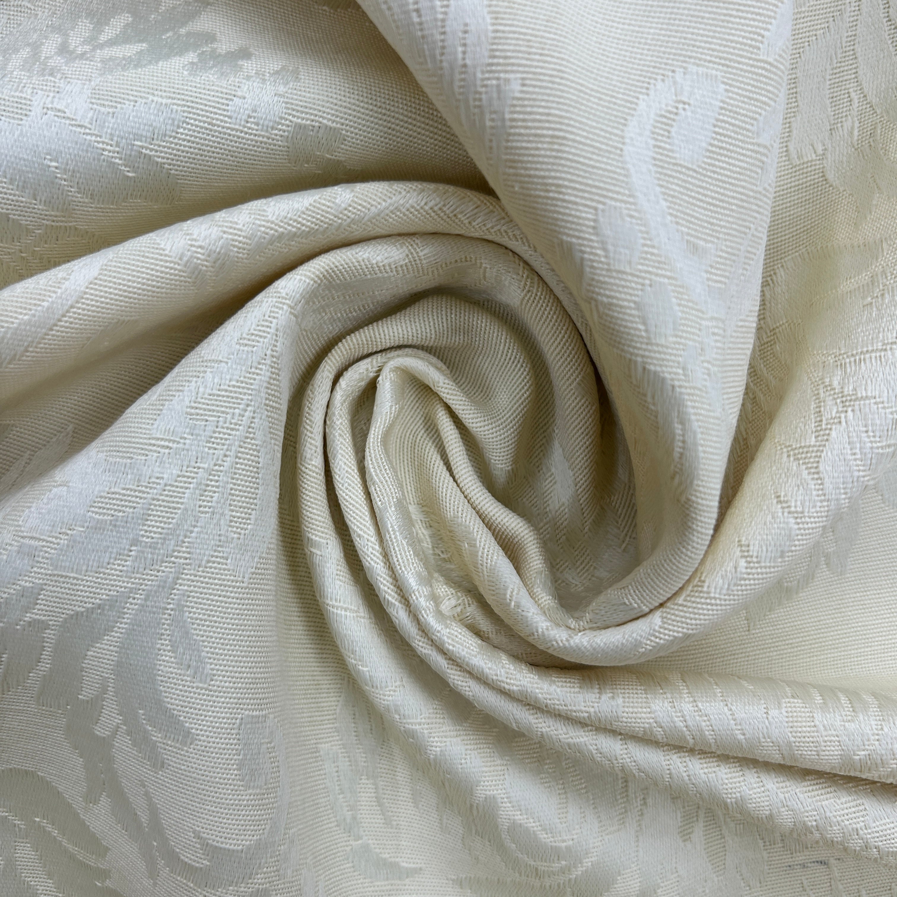 A close-up of draped cream-colored fabric with a subtle woven damask pattern, arranged in a spiral to show texture and sheen.