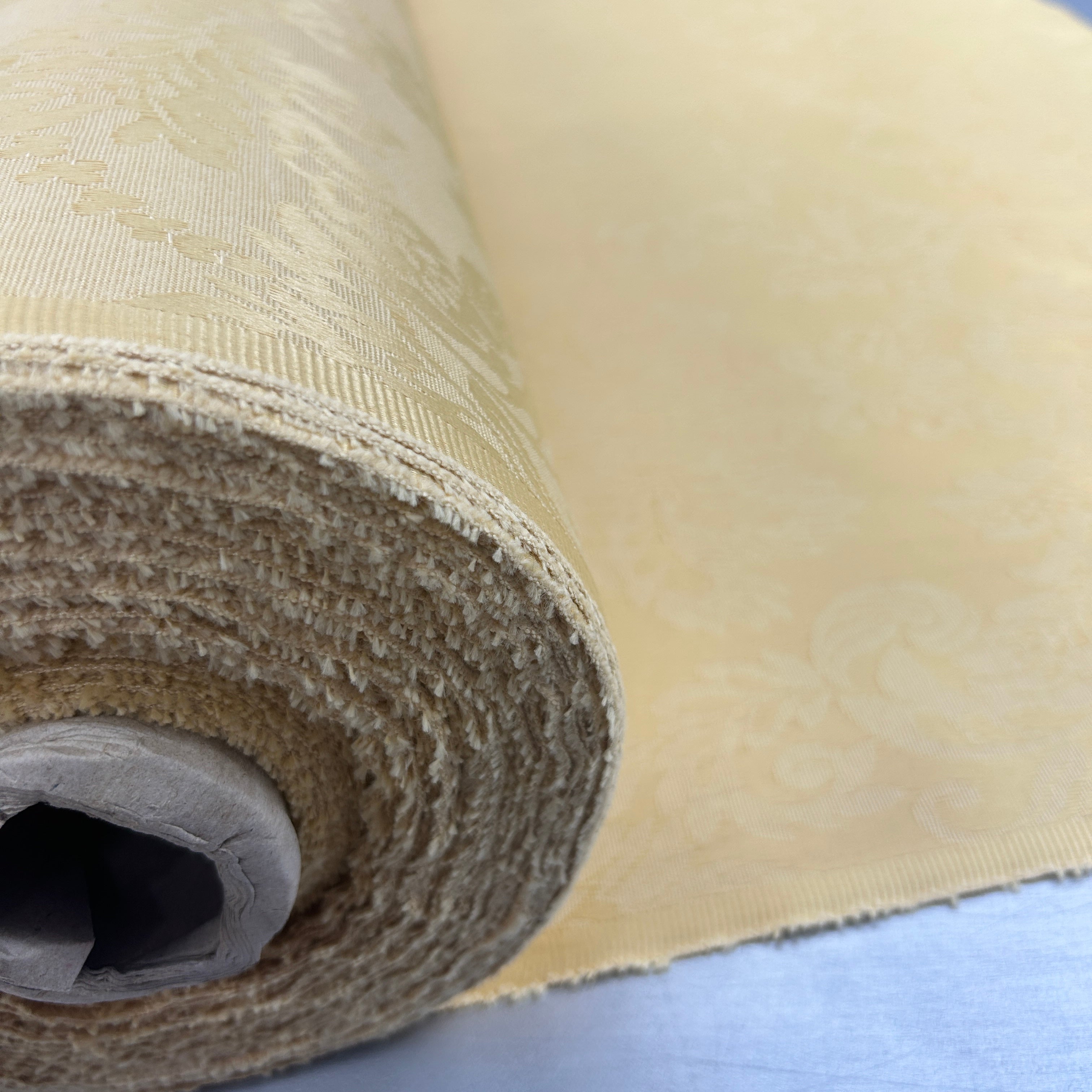 A close-up of a rolled beige fabric or wallpaper with a subtle damask pattern, showing both the textured rolled edge and the flat patterned surface.