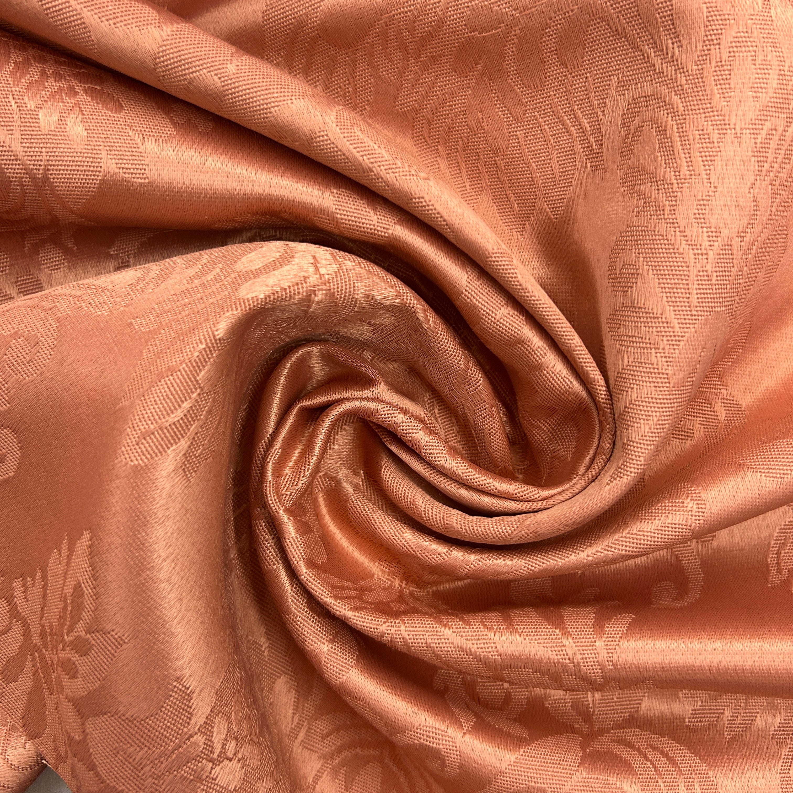 A close-up of peach-colored damask fabric with a subtle woven floral pattern, arranged in a spiral drape to show its texture and sheen.